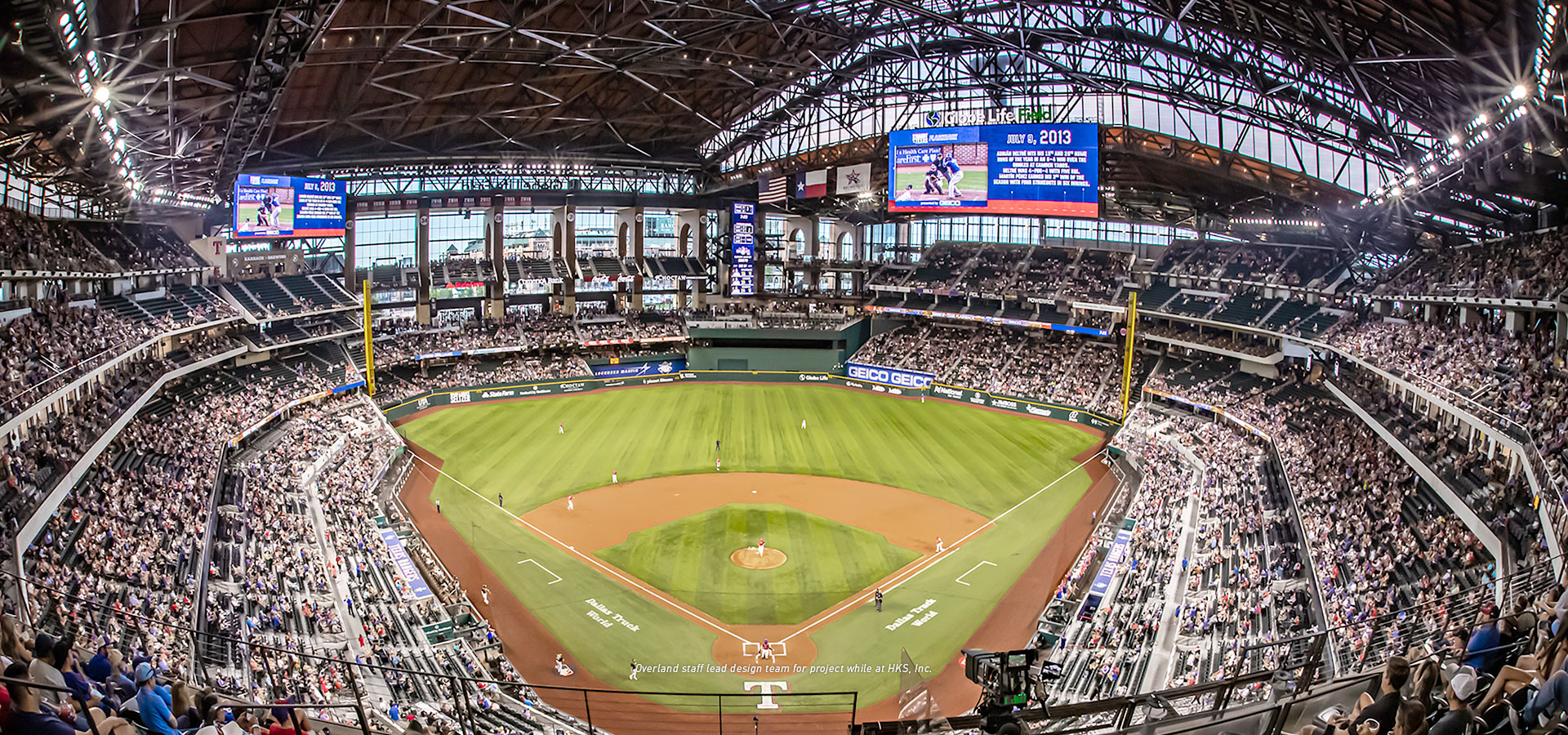 A large, covered stadium filled with spectators watching a baseball game. The field has a diamond layout with players positioned. The scoreboard at Globe Life Field is visible on the far side.