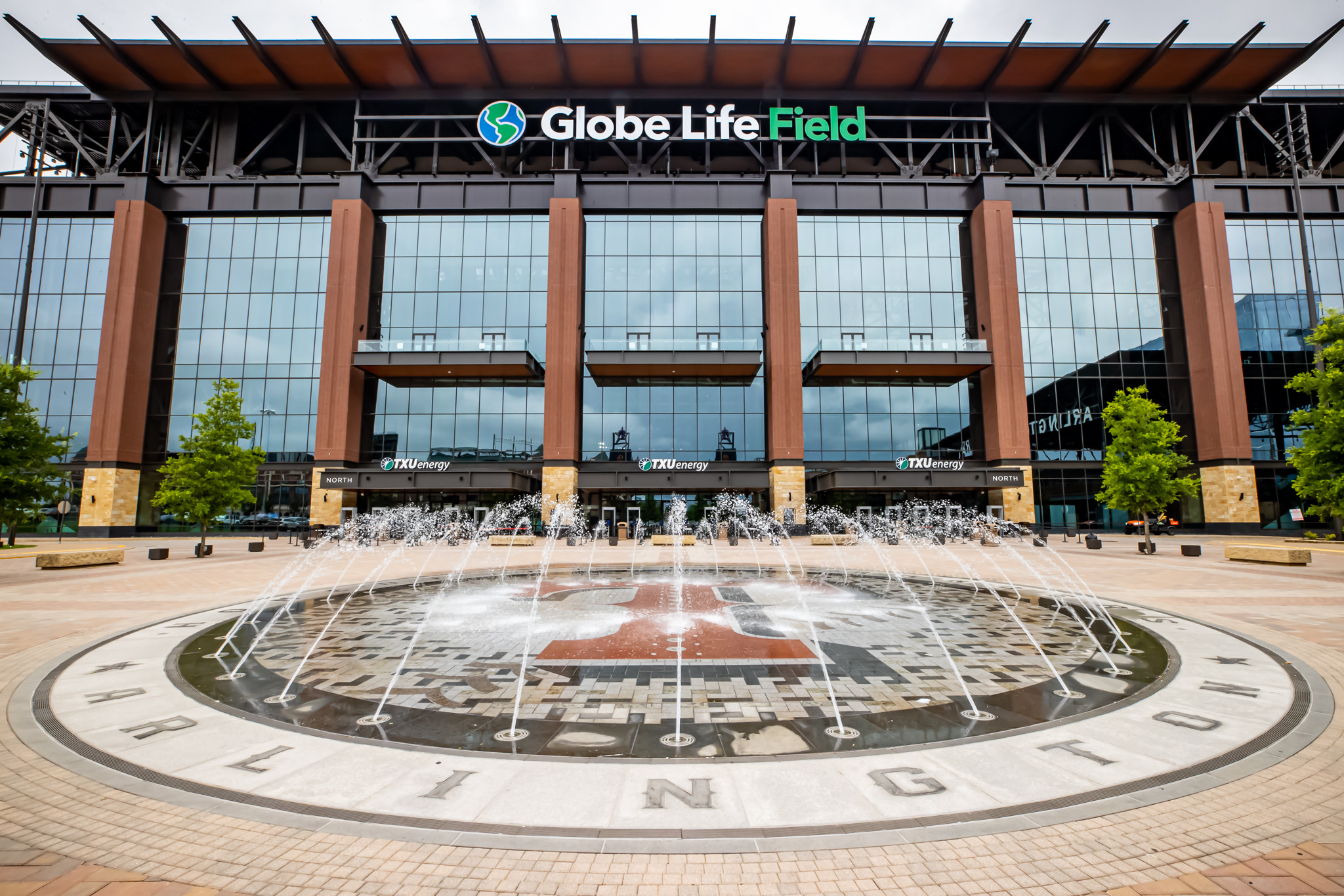 The exterior of Globe Life Field, with large glass windows, a prominent sign, and a water fountain in front, showcases the word "Arlington" on the pavement at this impressive sports venue.