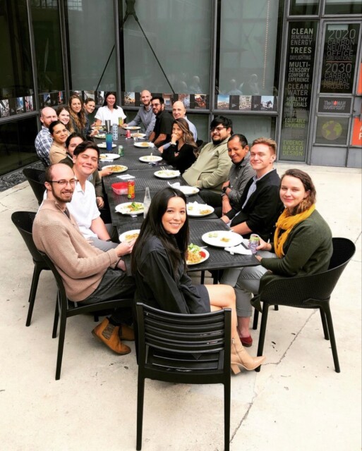 A group of people sitting at a long outdoor table, sharing a meal enriched with culture. Plates of food and drinks are in front of them. They are smiling and looking towards the camera, with indoor plants visible in the background.