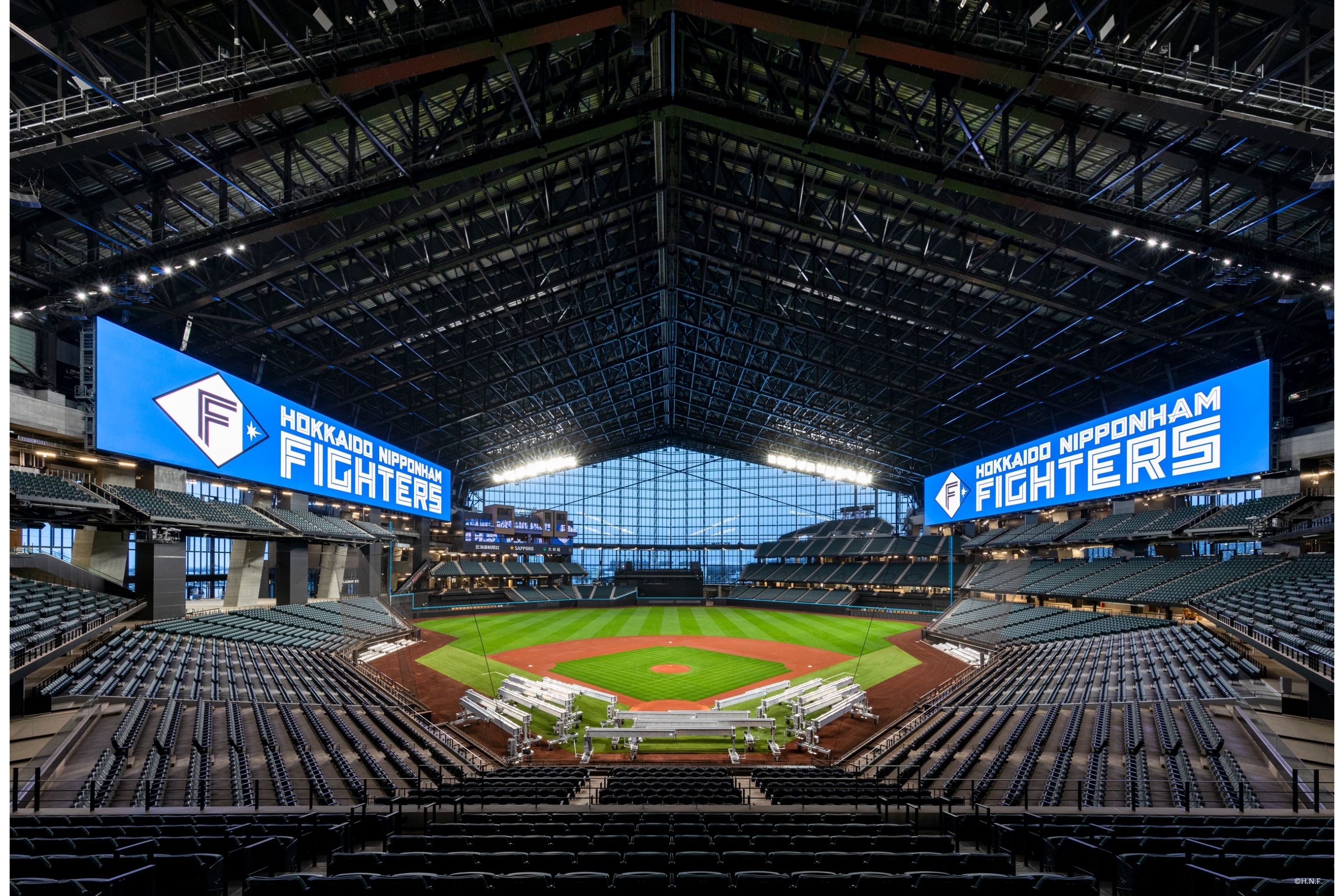 An empty indoor baseball stadium with a large roof and seating. Two large screens display "Hokkaido Nippon-Ham Fighters." The well-lit and neatly maintained baseball field showcases the impeccable standards of ES Con Field Hokkaido.