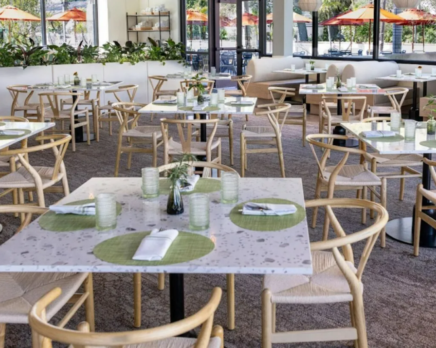 A brightly lit net-positive restaurant with light wooden chairs and white tables set with green placemats, glasses, napkins, and small plants. Outside the windows are patio seats with orange umbrellas, reminiscent of the vibrant flora at Selby Gardens in Florida.