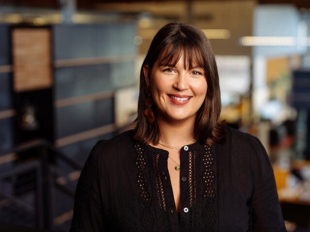 Shelby Sekaly, a woman with shoulder-length brown hair and a black blouse, smiles in an office setting with a blurred background.