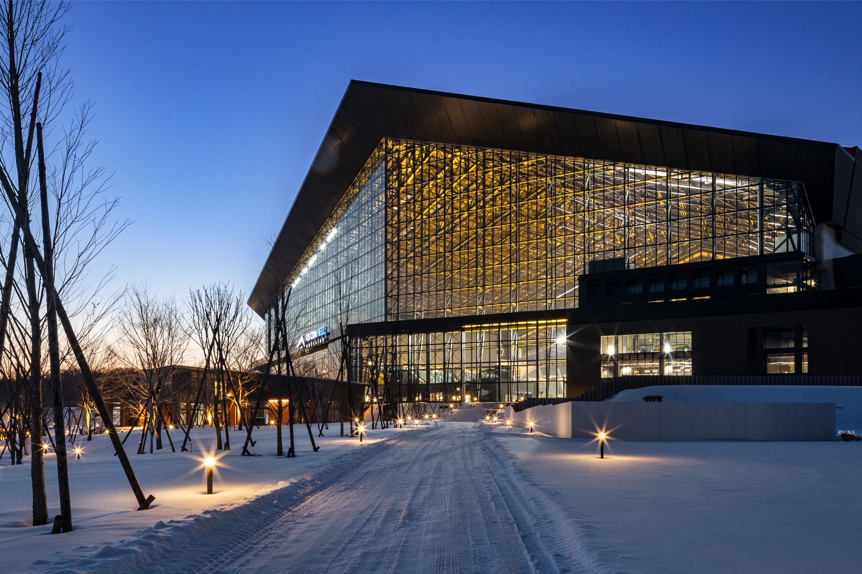 A modern building with large, illuminated glass walls stands amid a snowy landscape at dusk in Hokkaido. Bare trees line the path leading to the entrance of ES Con Field, highlighted by ground lights.