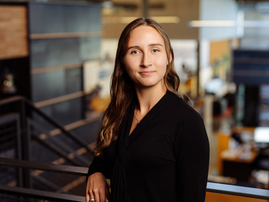 A woman with long brown hair, wearing a black blouse, stands in a modern office environment, facing the camera and smiling slightly. Samantha Braswell exudes confidence as she navigates her professional surroundings.