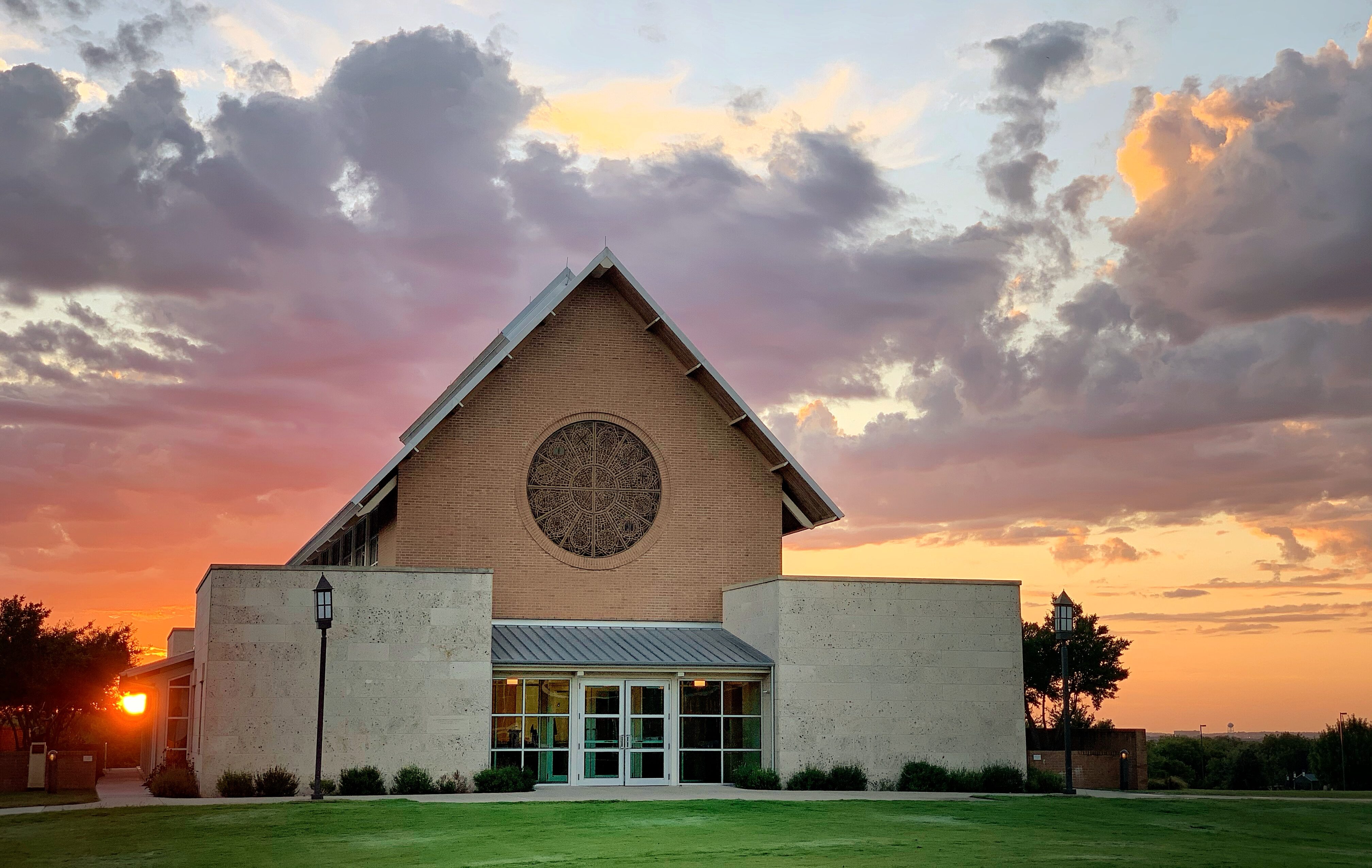 Against a colorful sunset sky with clouds, the church building stands majestically, its large circular window resembling an artist's auto draft of divine beauty.