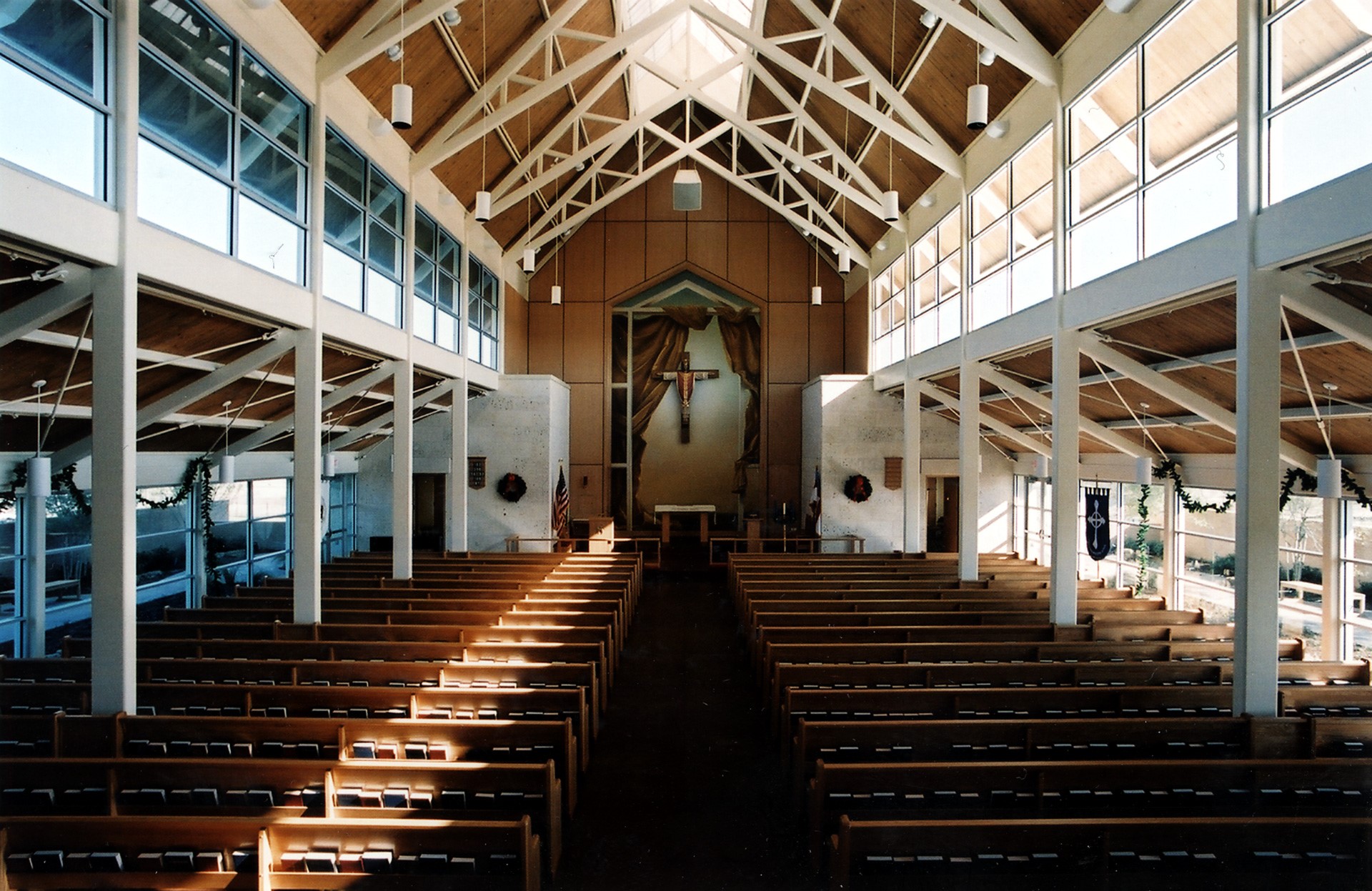 Inside this modern church, wooden pews line the nave beneath a ceiling of exposed beams. Soft light filters through expansive windows, casting a serene glow on the cross at the front. The architecture invites tranquility and reflection, creating an inspiring space for worship and contemplation.