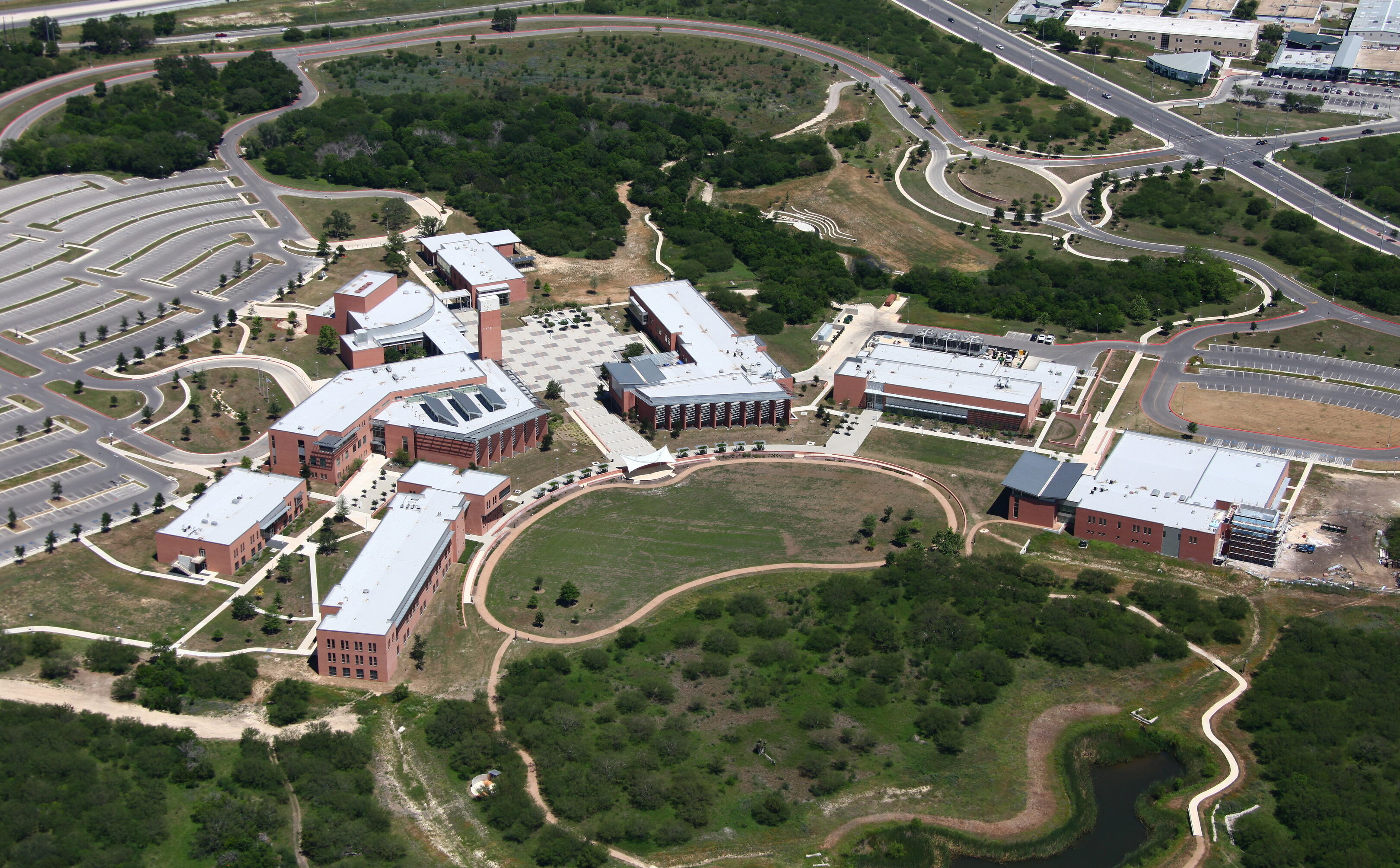 Aerial view of a vibrant campus with red-brick buildings, lush green spaces, and well-organized parking lots. Intersecting roads and pathways weave through the site, creating a picturesque blend of nature and architecture.