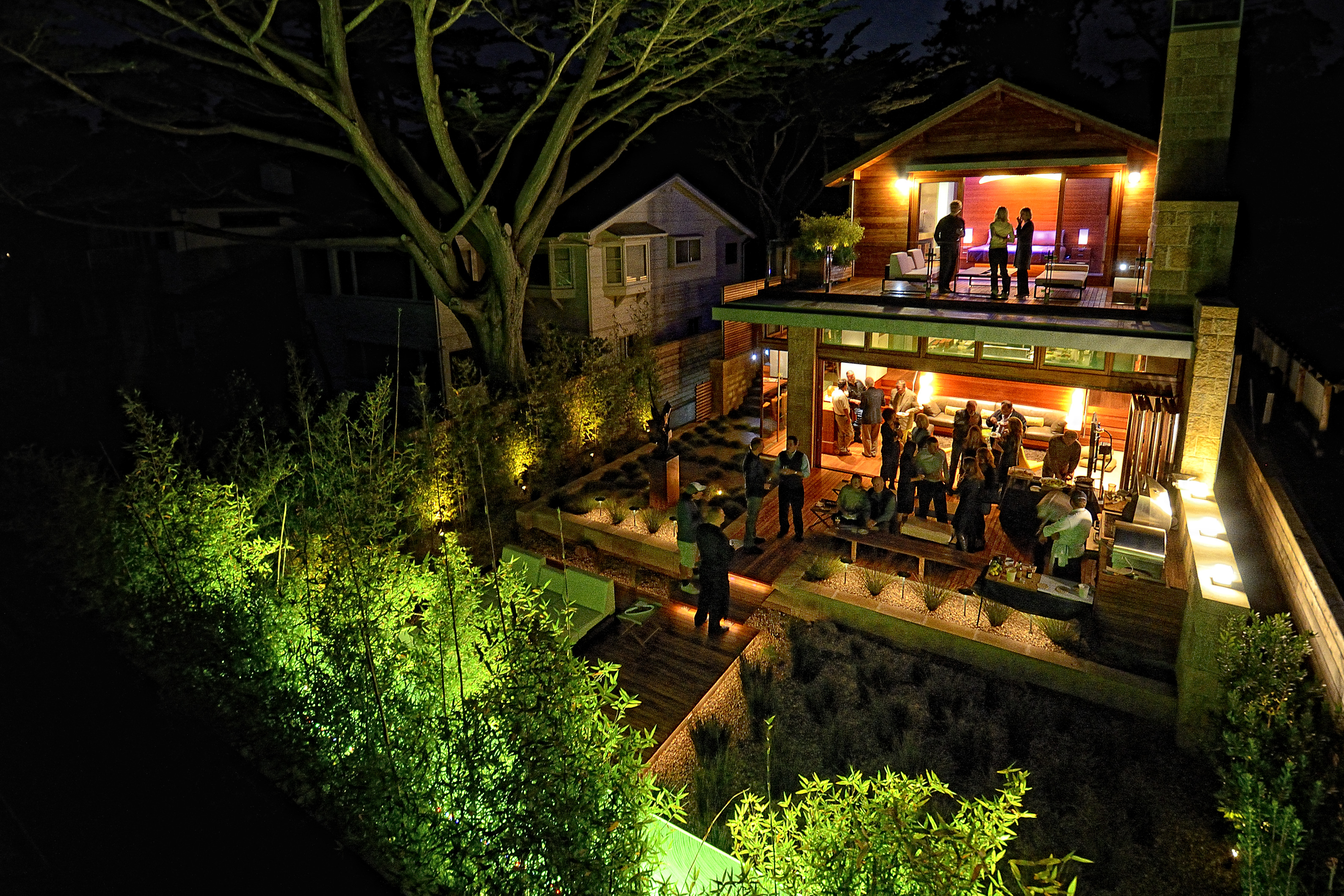 At the Carmel Residence, a nighttime backyard party unfolds like an odyssey, with guests gathered on a lit patio and upstairs balcony, surrounded by illuminated trees and vibrant plants.