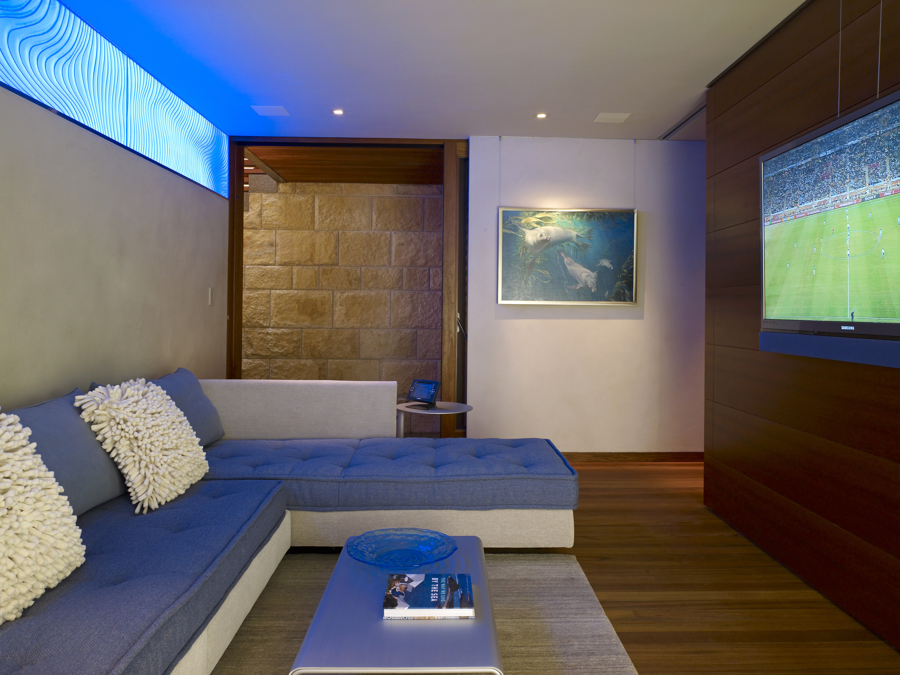 Modern living room in the Carmel residence, featuring a gray sectional couch, cozy cushions, and a wall-mounted TV showing a sports game. Blue lighting glows above, enhancing the ambiance, while an ocean-themed picture frames an artistic odyssey on the wall.