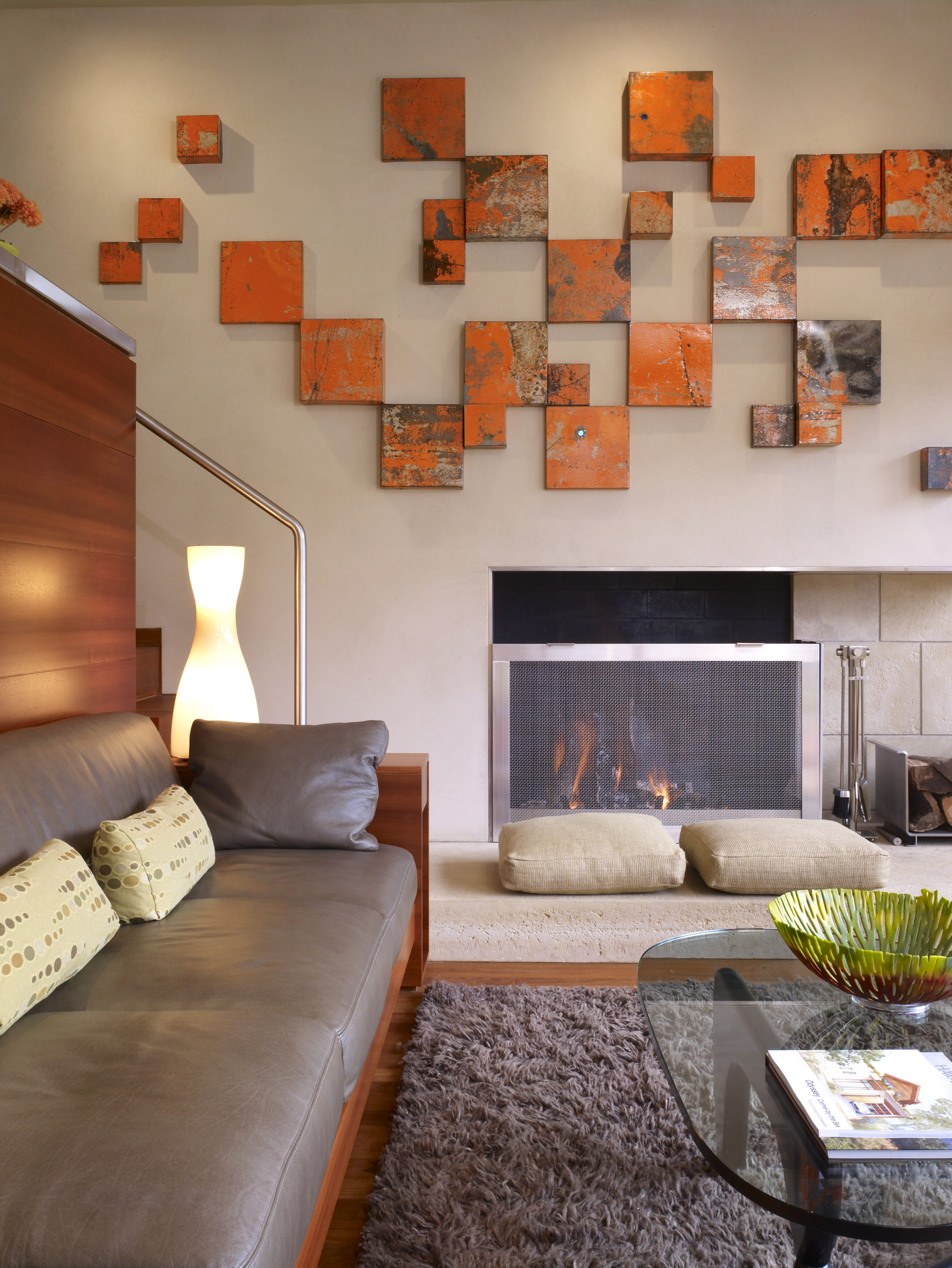 A modern living room in the Carmel residence features a gray couch, a lit lamp, and a cozy fireplace. Abstract orange art hangs above, adding warmth to the space. A glass coffee table and textured rug complete this stylish odyssey of contemporary decor.