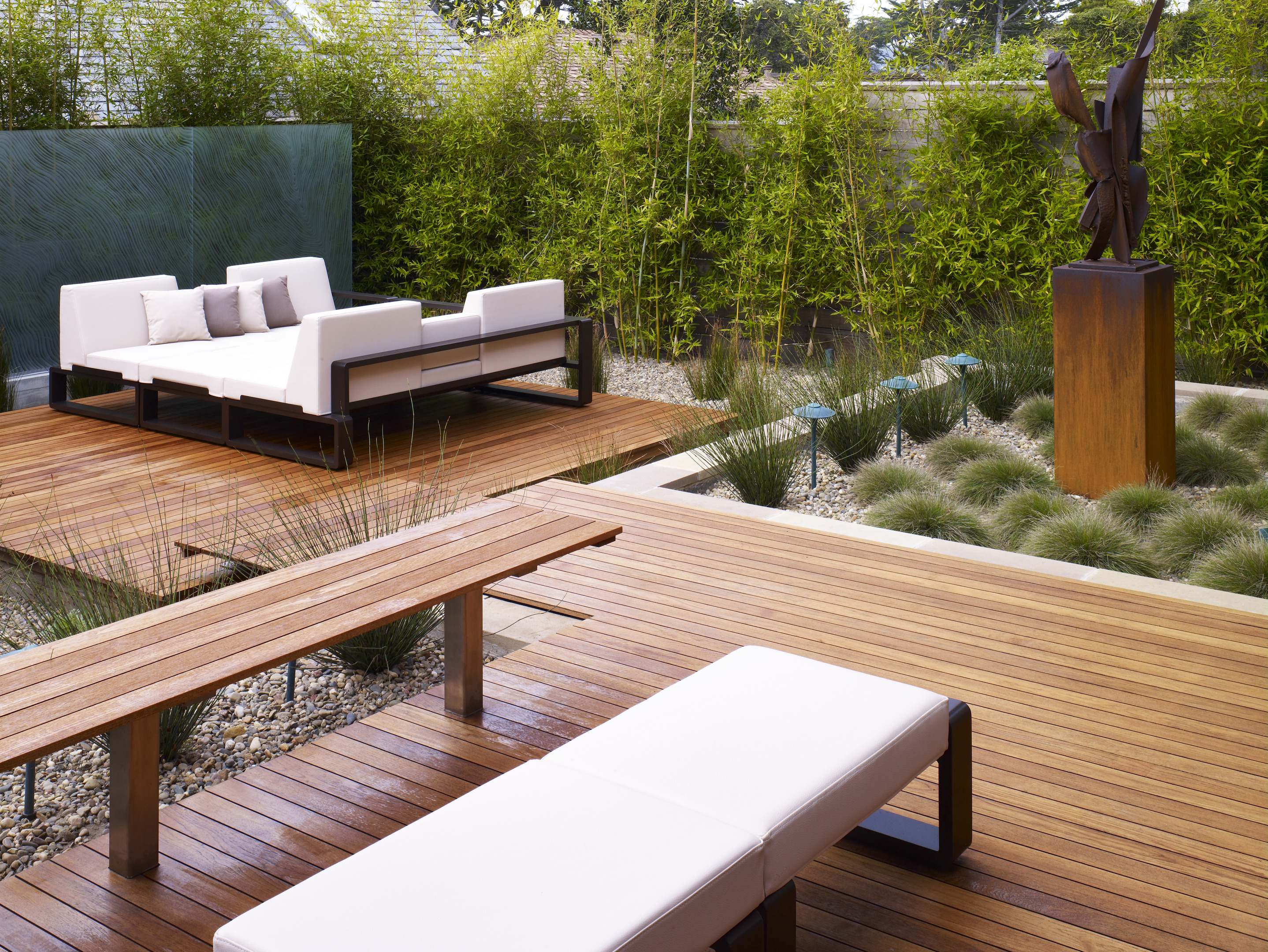 The Carmel Residence patio features white seating on wooden decking, surrounded by decorative grasses and a large abstract sculpture. Bamboo plants create a green backdrop, adding an odyssey of peaceful retreat to the modern design.