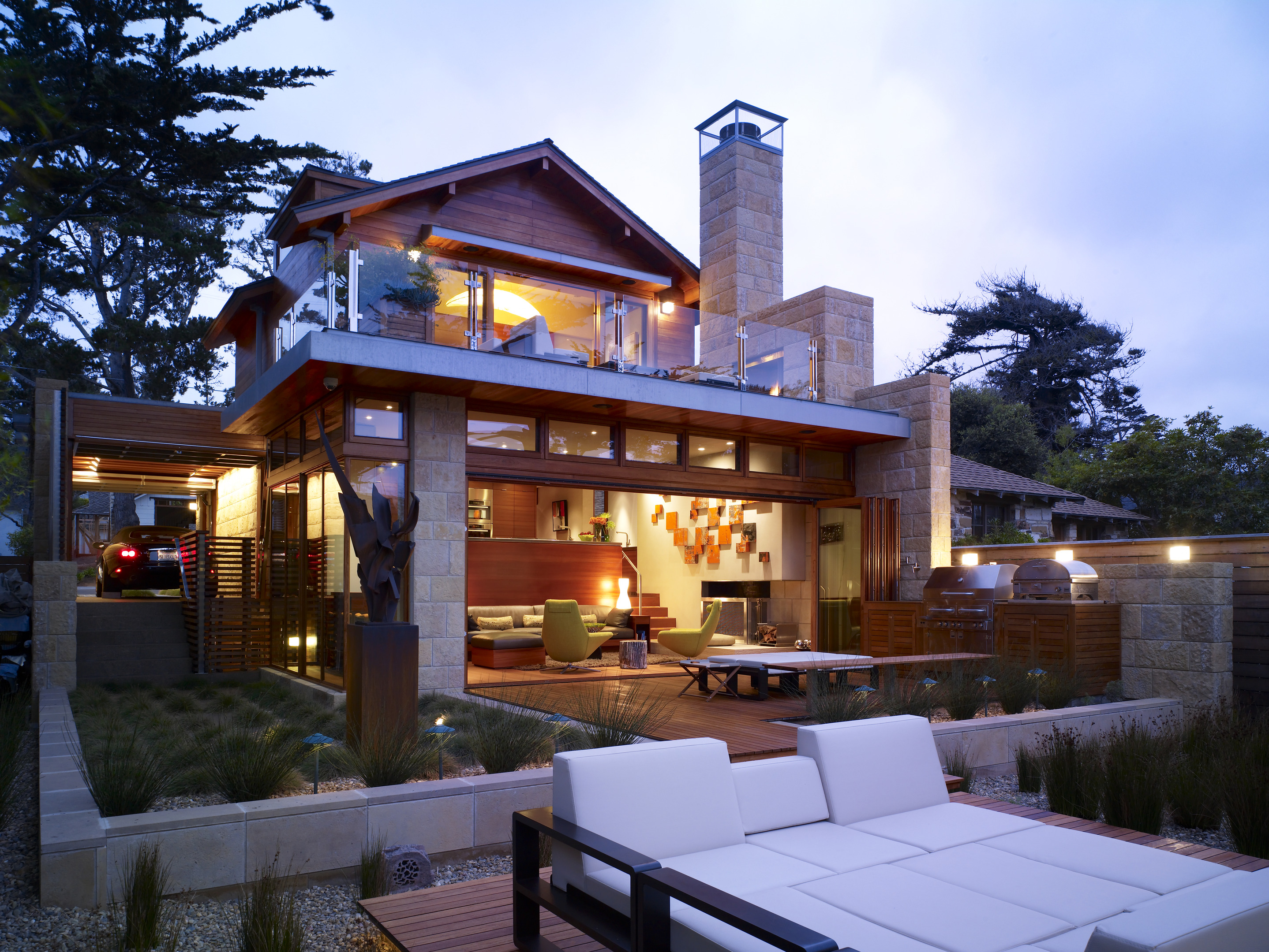 The Carmel Residence is a modern multi-level house with large windows, a rooftop sitting area, and a deck filled with outdoor furniture. Its illuminated interior glows through the glass, nestled amidst trees and greenery. An architectural odyssey in harmony with nature.