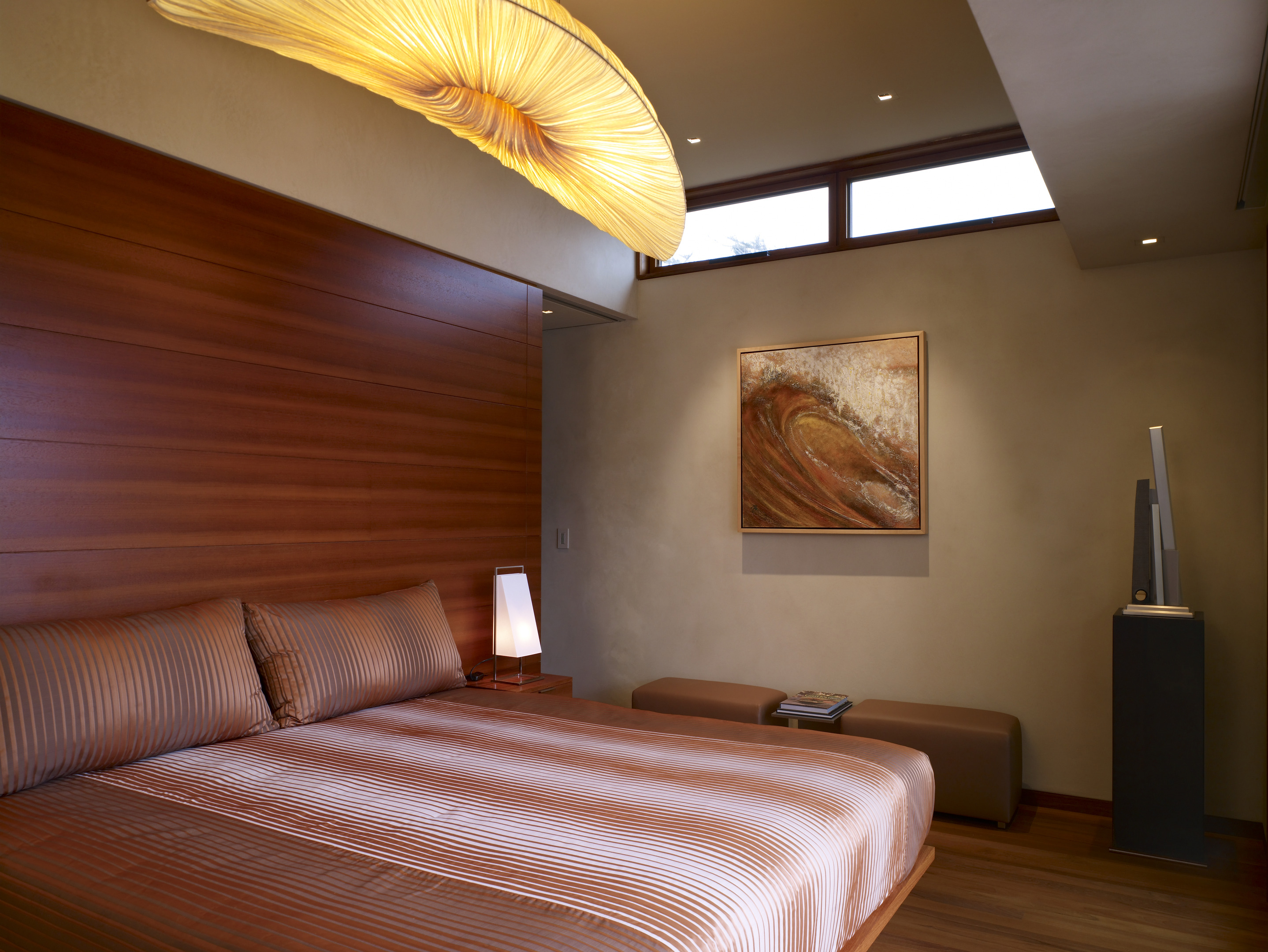 A modern bedroom in the Carmel Residence features a large bed, wooden accents, a unique ceiling light, a wall painting, a small lamp, and a bench beside a flat-screen TV—an interior design odyssey.