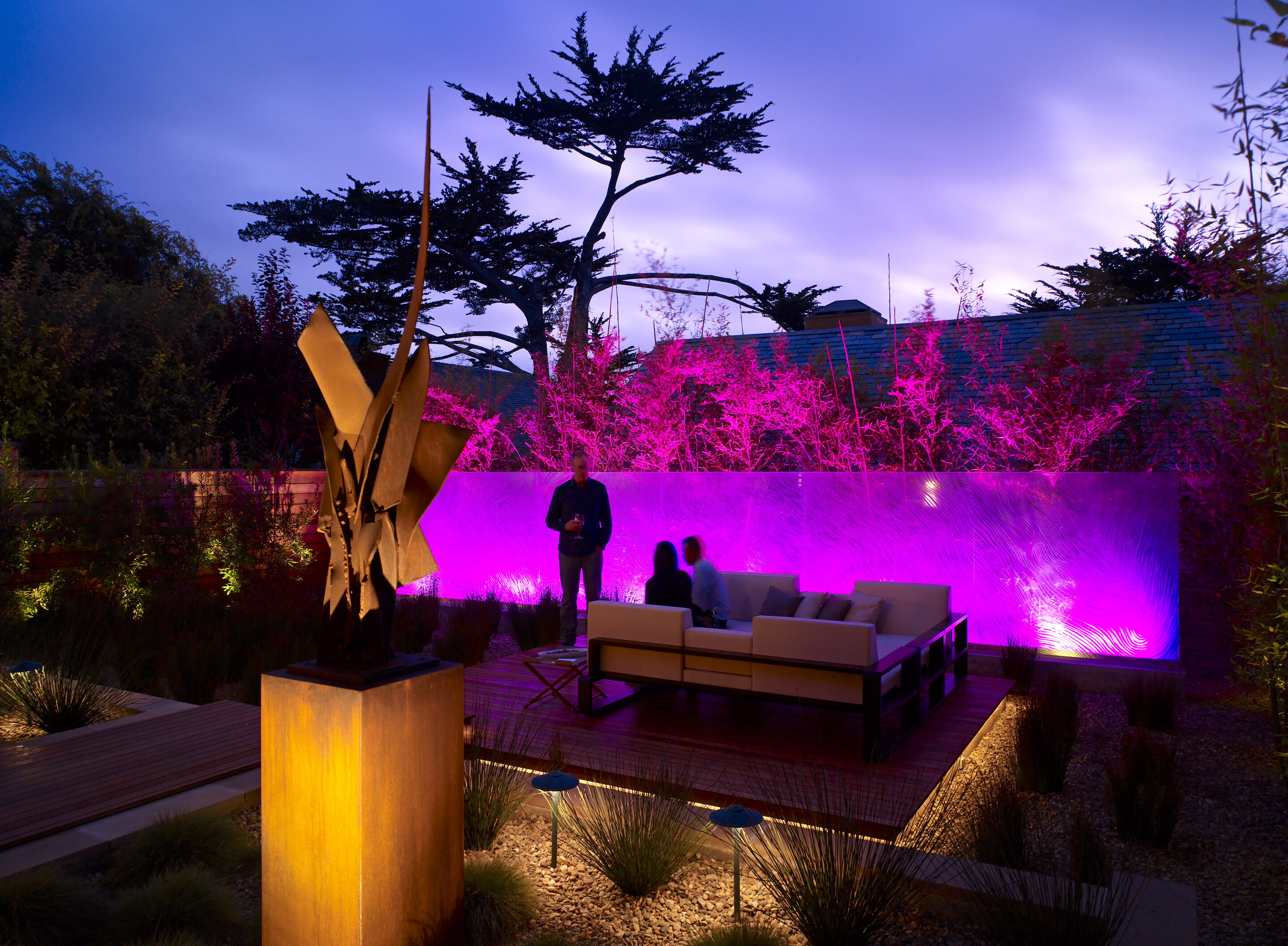 The outdoor seating area at Carmel Residence features a modern sculpture, illuminated by pink and purple lights. Trees sway against the backdrop of a cloudy sky, creating an odyssey of art and nature.