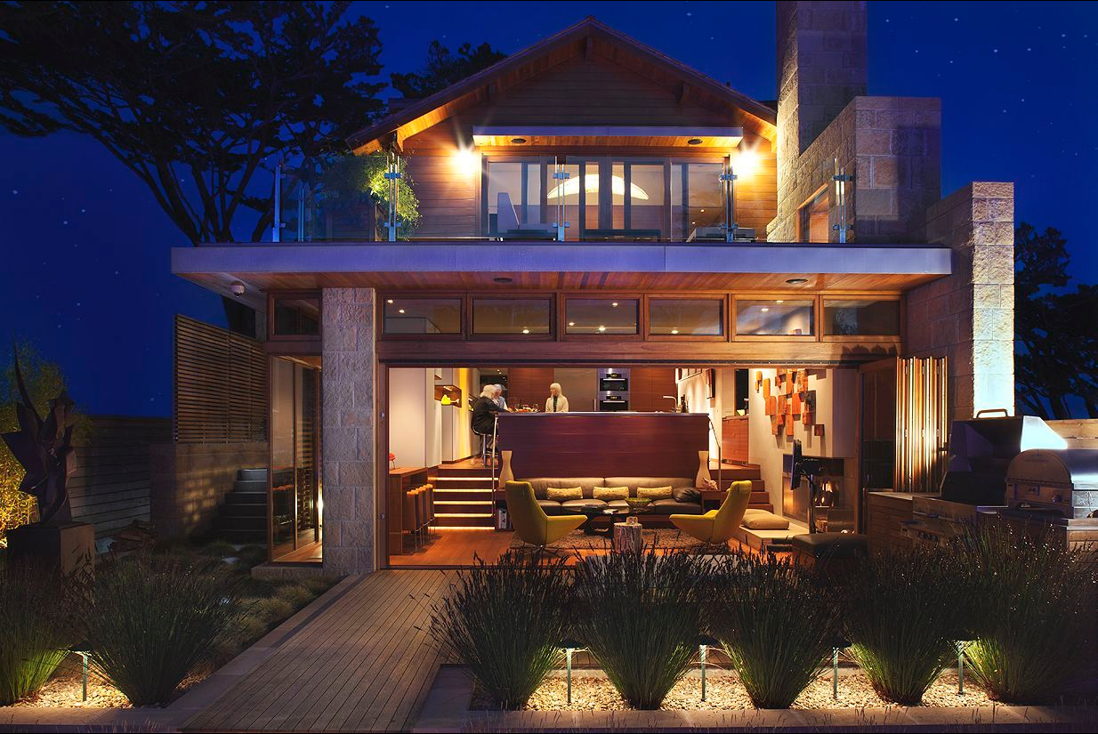 Modern two-story residence illuminated at night, featuring large windows, a wood and stone exterior, and a landscaped garden reminiscent of an architectural odyssey.