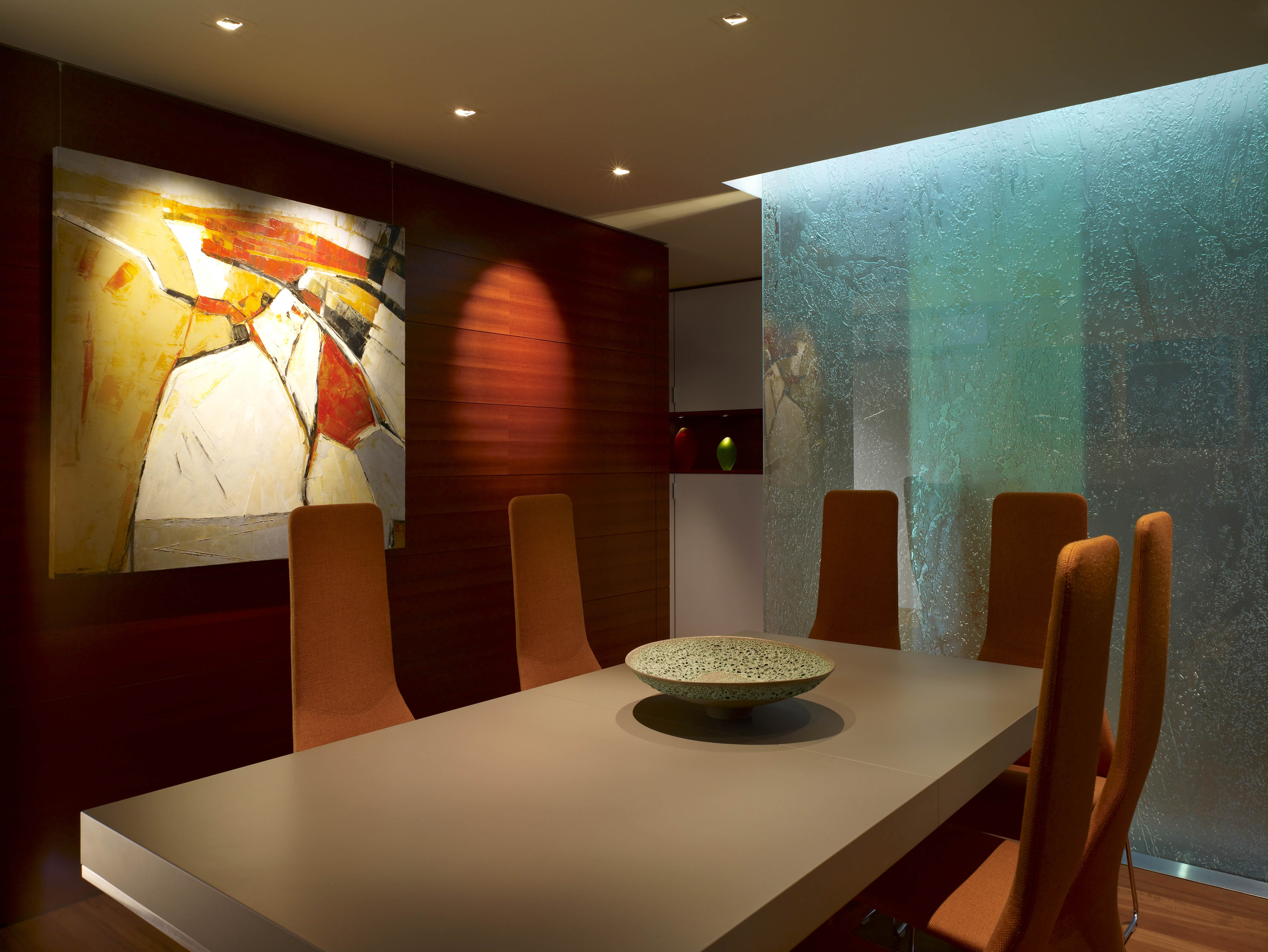 The Carmel Residence features a modern dining room with a rectangular table, six vibrant orange chairs, an abstract painting adorning the wood-paneled wall, and a textured glass partition that completes this artistic odyssey of design.