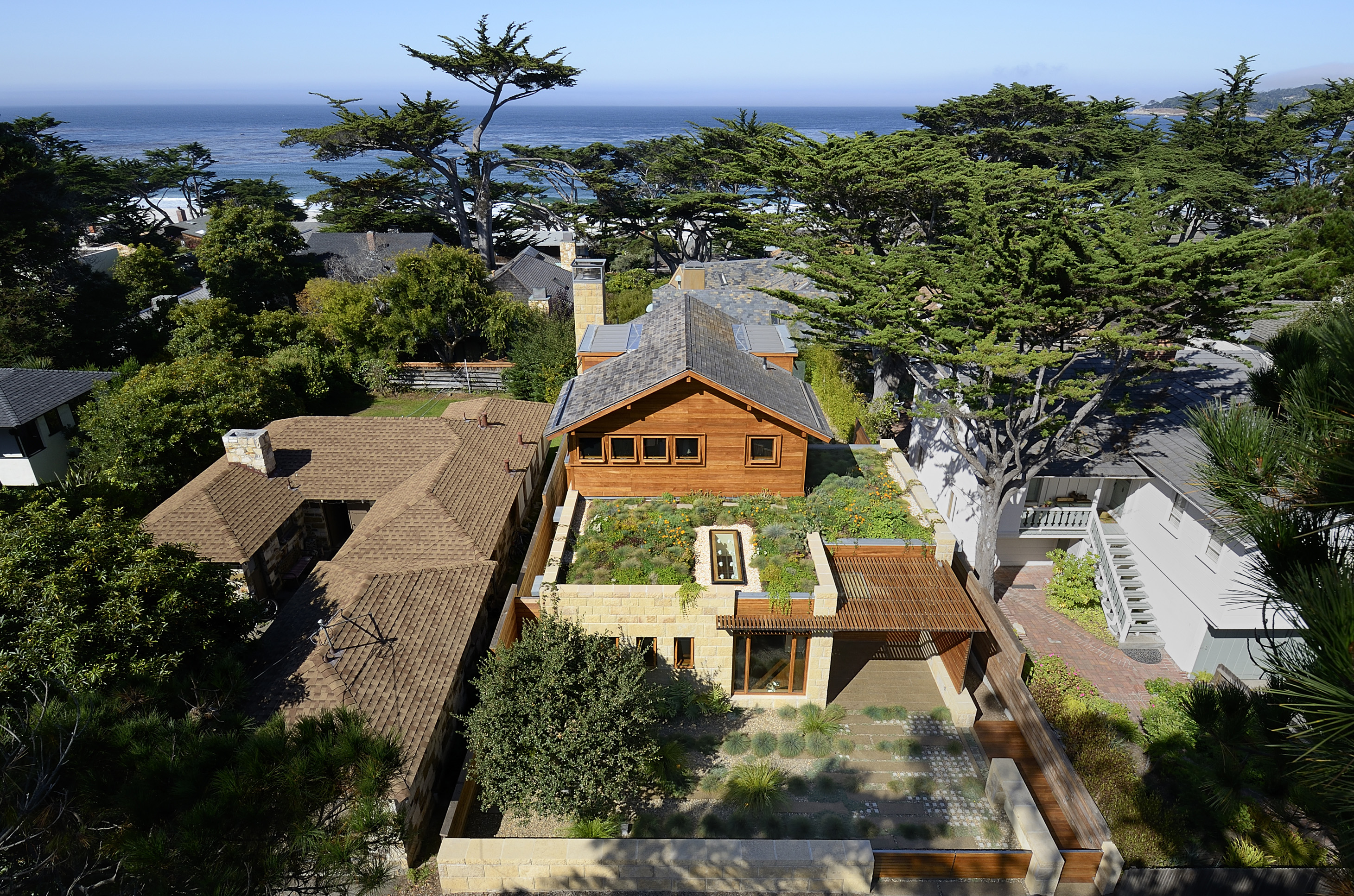 Aerial view of the Carmel residence, a modern wooden house with a rooftop garden, nestled among trees and other homes near the ocean.