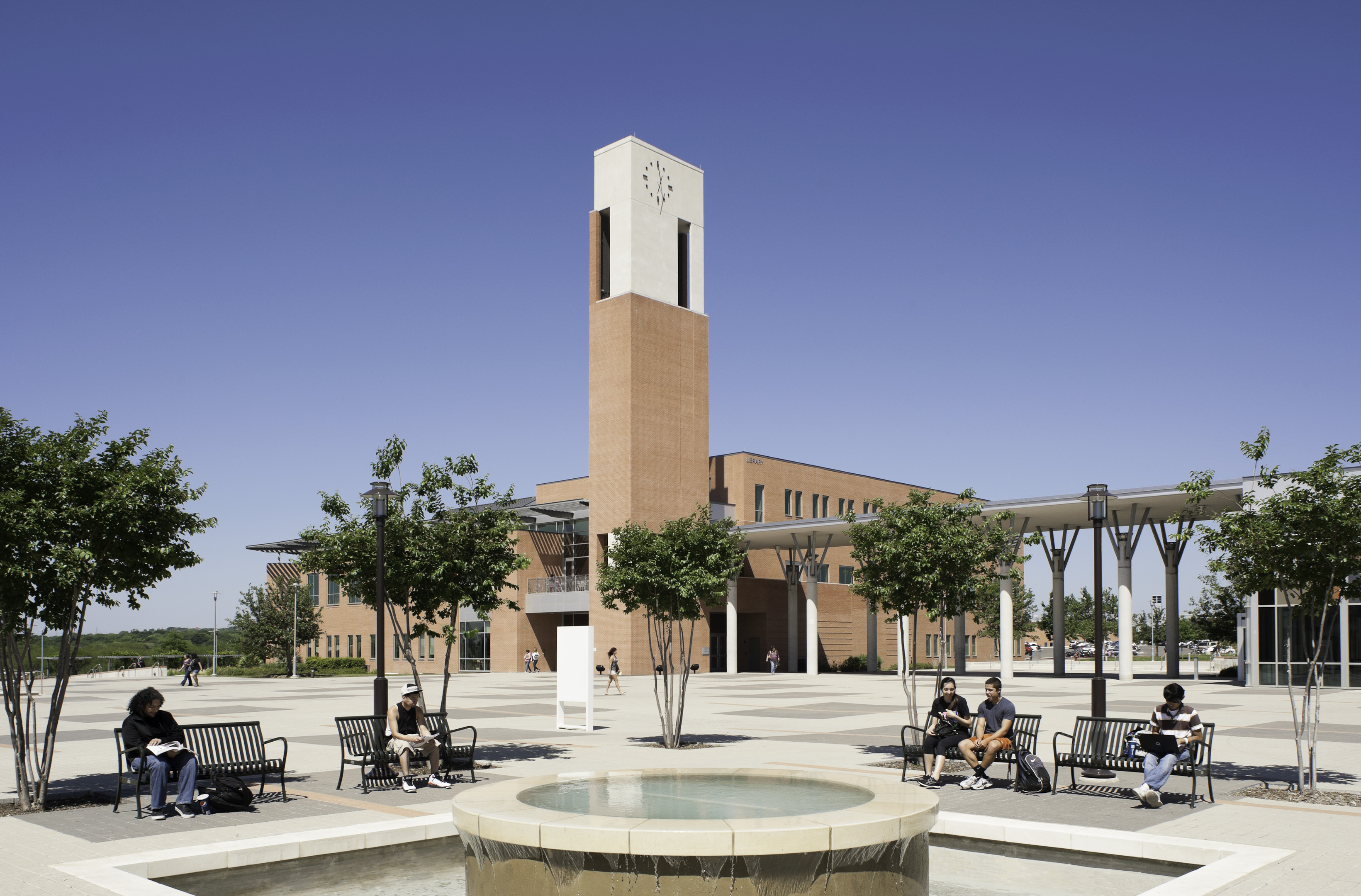 A picturesque university campus features a classic brick clock tower, where students enjoy leisurely moments on benches amid lush trees and a fountain under the expansive, clear blue sky.