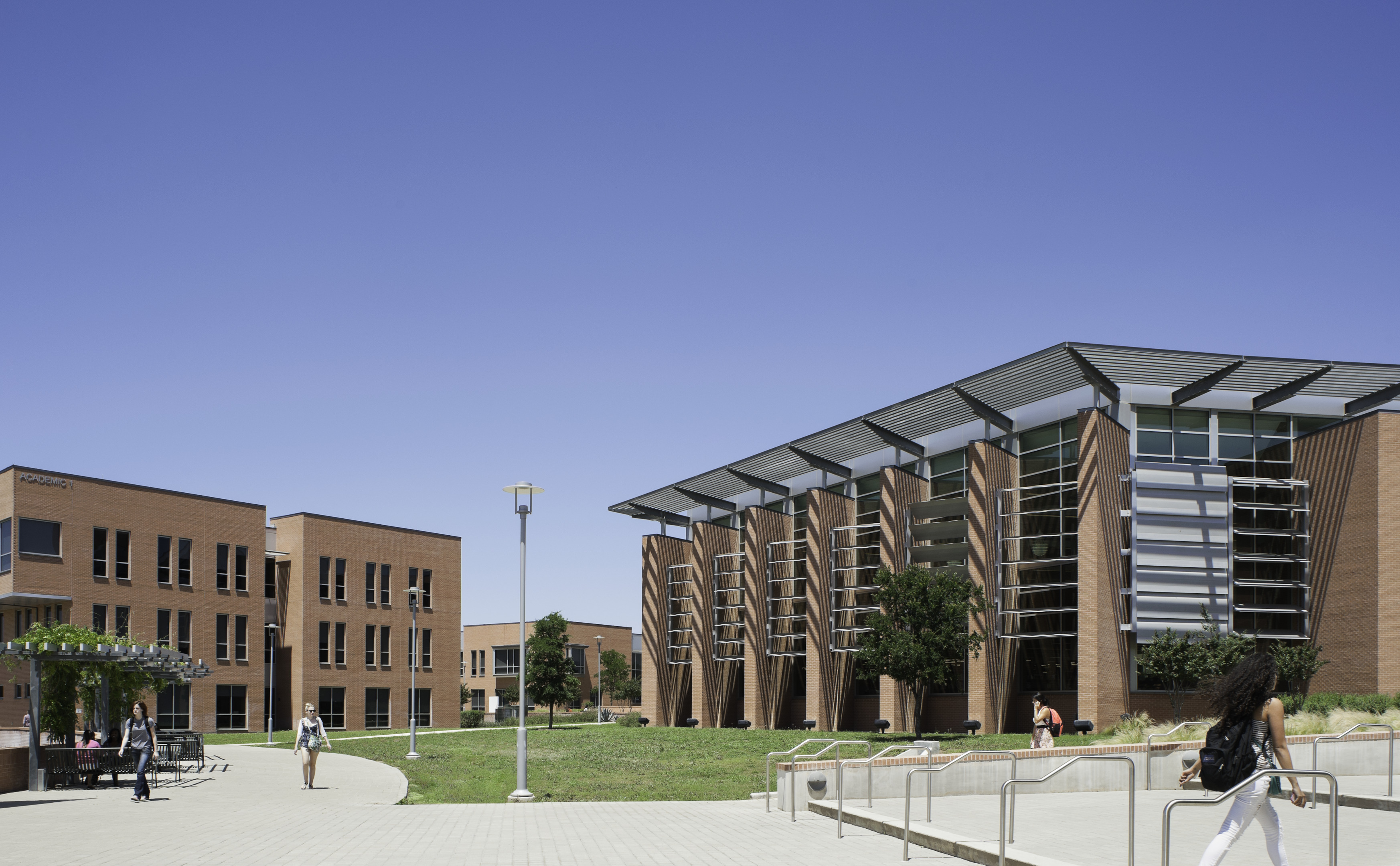 The modern university campus, with two brick buildings under a clear blue sky, resembles an auto draft of architectural harmony, as a few people casually stroll along the pathways.