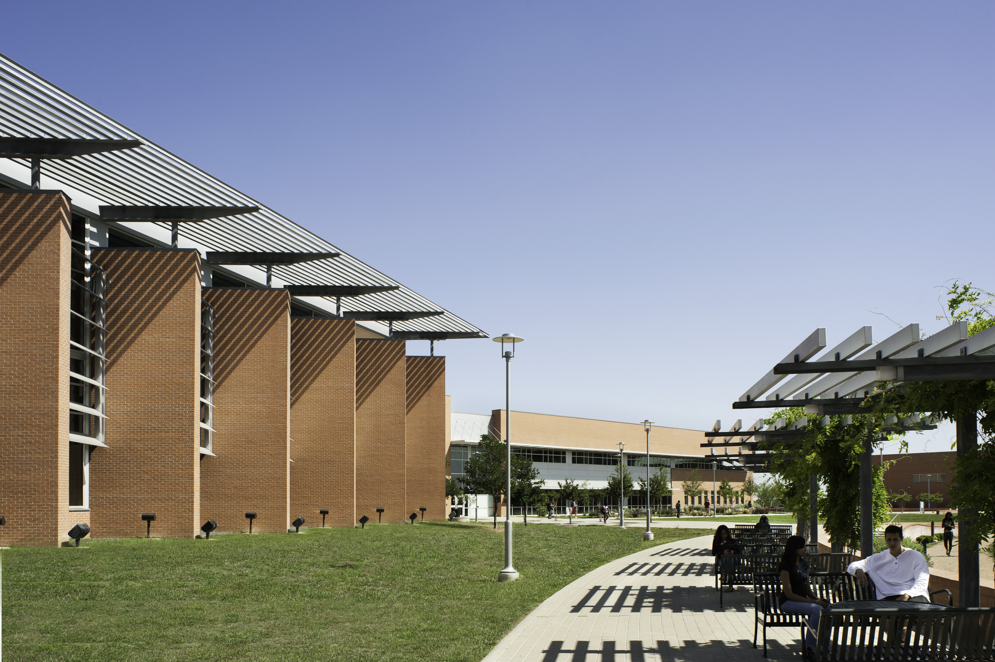 Brick building with a modern design, highlighting tall windows and a shaded outdoor pathway. Under the pergola, people enjoy the scenery while seated on benches. With a clear blue sky in the background, this scene feels almost like an auto draft of architectural perfection.