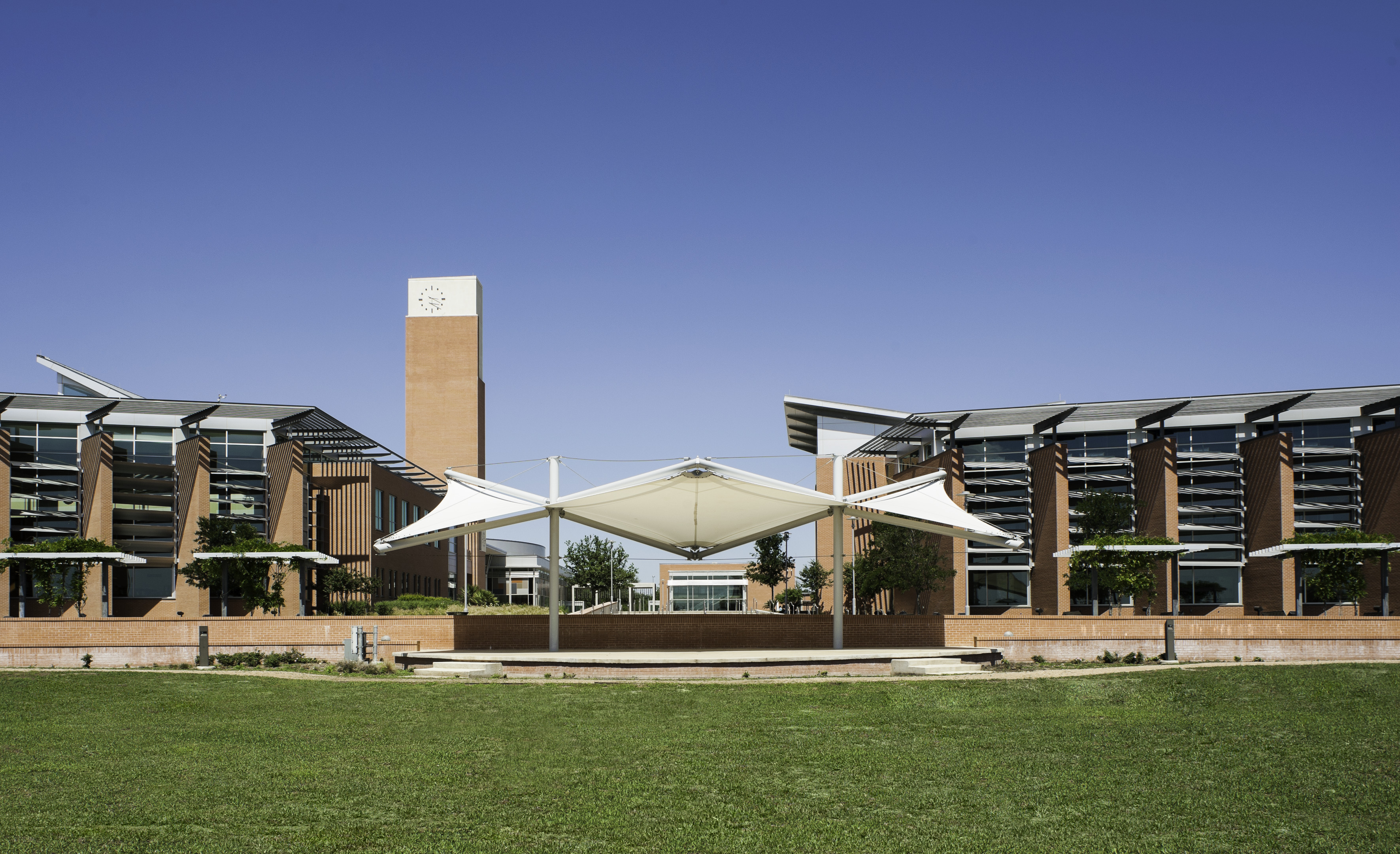 A modern building complex with a central canopy structure, lush grassy foreground, and a clock tower rising in the background under a clear blue sky, creating an atmosphere reminiscent of an auto draft masterpiece.