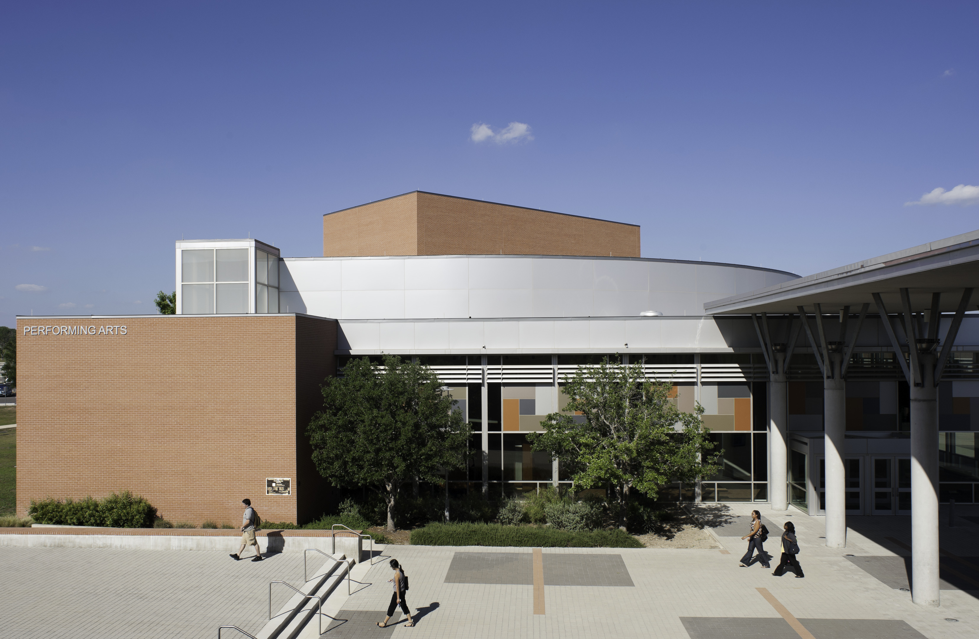 A modern performing arts center with an auto draft-inspired geometric design features a light brown and silver facade. Under the clear blue sky, three people stroll across the plaza, adding life to the architectural masterpiece.