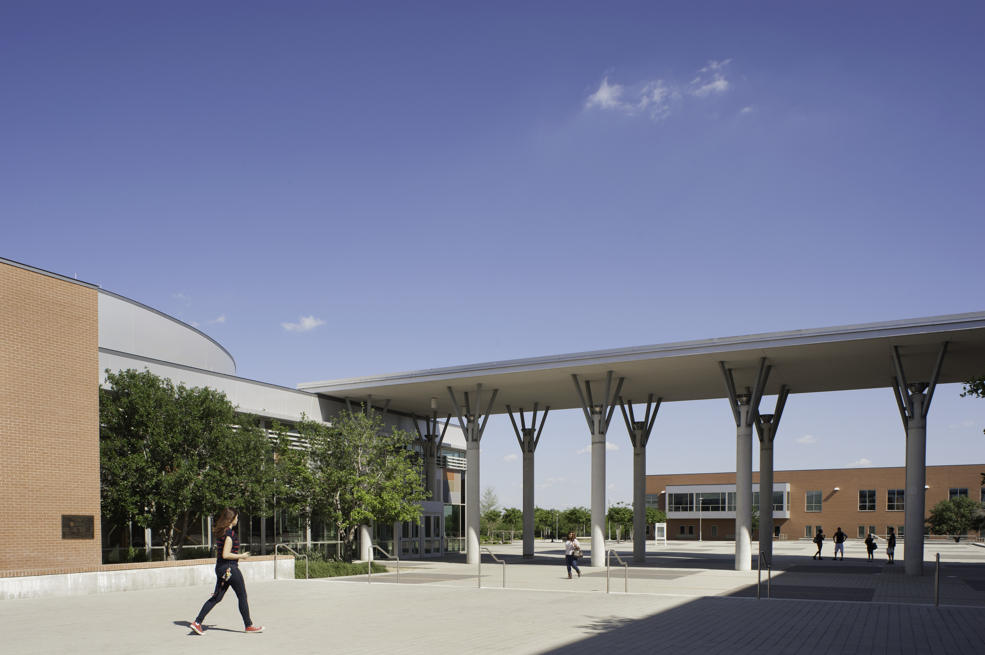 People walk across a courtyard between modern brick buildings under a clear blue sky, as if entering an auto-drafted scene in an architect's vision.