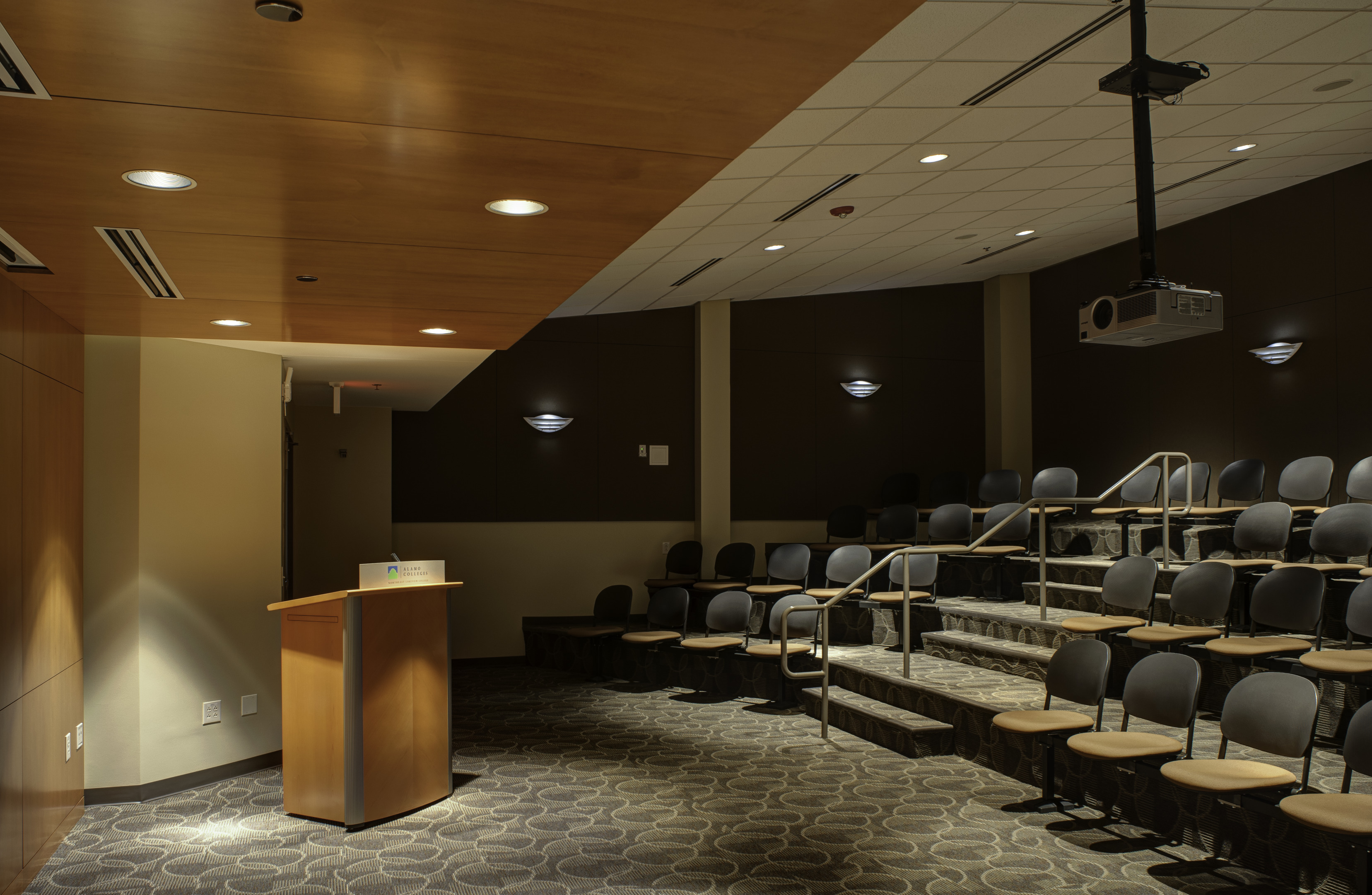 An empty lecture hall with tiered seating, a podium, and an overhead projector stands ready, its carpeted flooring echoing the anticipation of future discussions just waiting for the auto-draft of brilliant ideas.