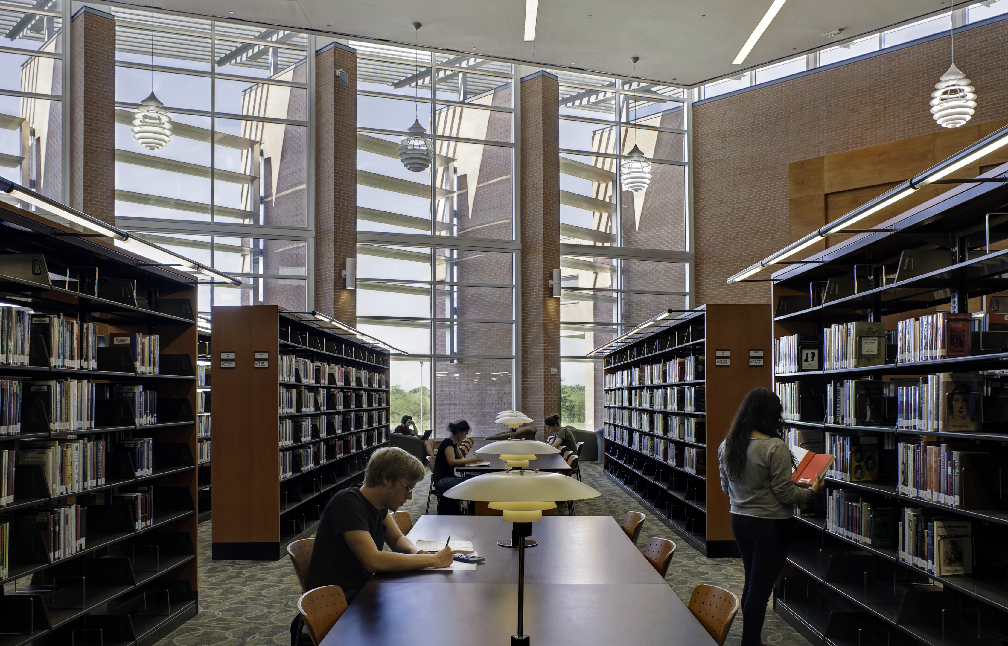 In a spacious library with high ceilings and large windows, people are immersed in reading and studying, surrounded by bookshelves on both sides—a serene environment that almost feels as if words gracefully auto-draft themselves into their thoughts.