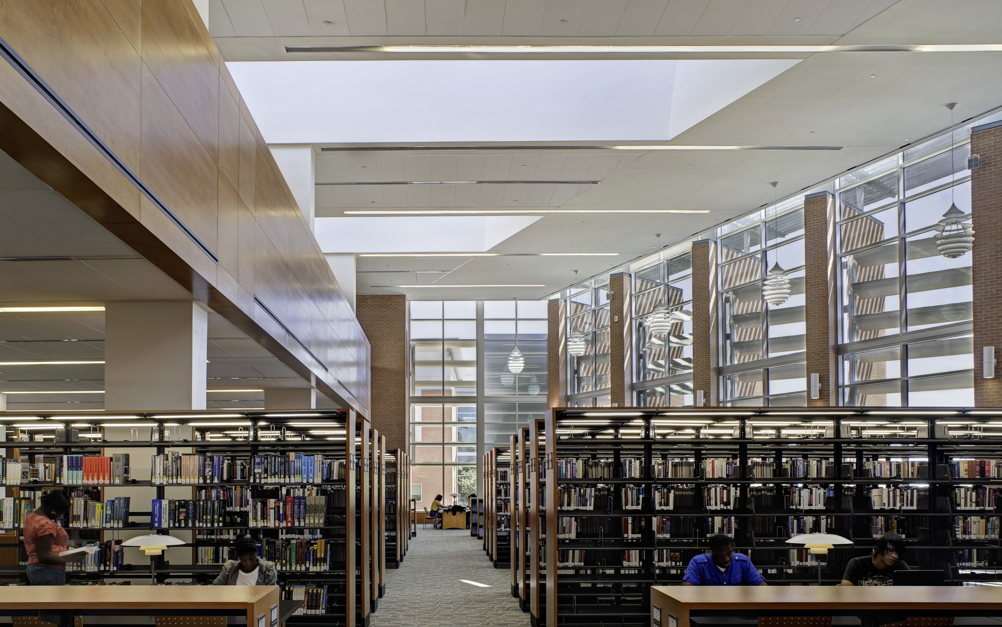 The interior of a modern library with tall glass windows and bookshelves creates an inspiring backdrop as people study at desks, their laptops open like an auto draft, effortlessly capturing the flow of ideas.