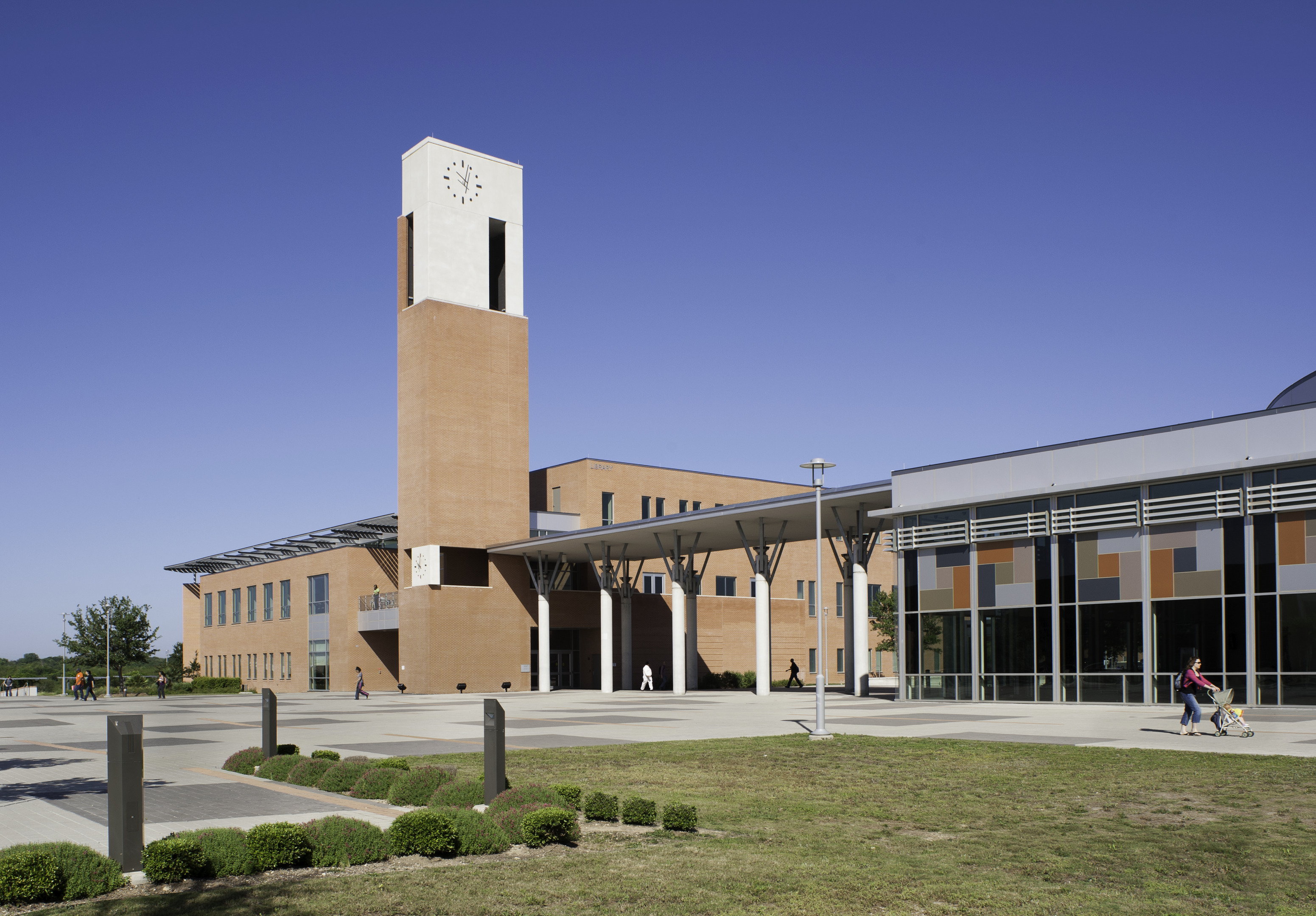 The modern building, featuring a tall clock tower and large windows, stands majestically with its spacious outdoor area under a clear blue sky. Nearby, people are walking and cycling as if part of an urban auto draft, seamlessly blending into the vibrant cityscape.