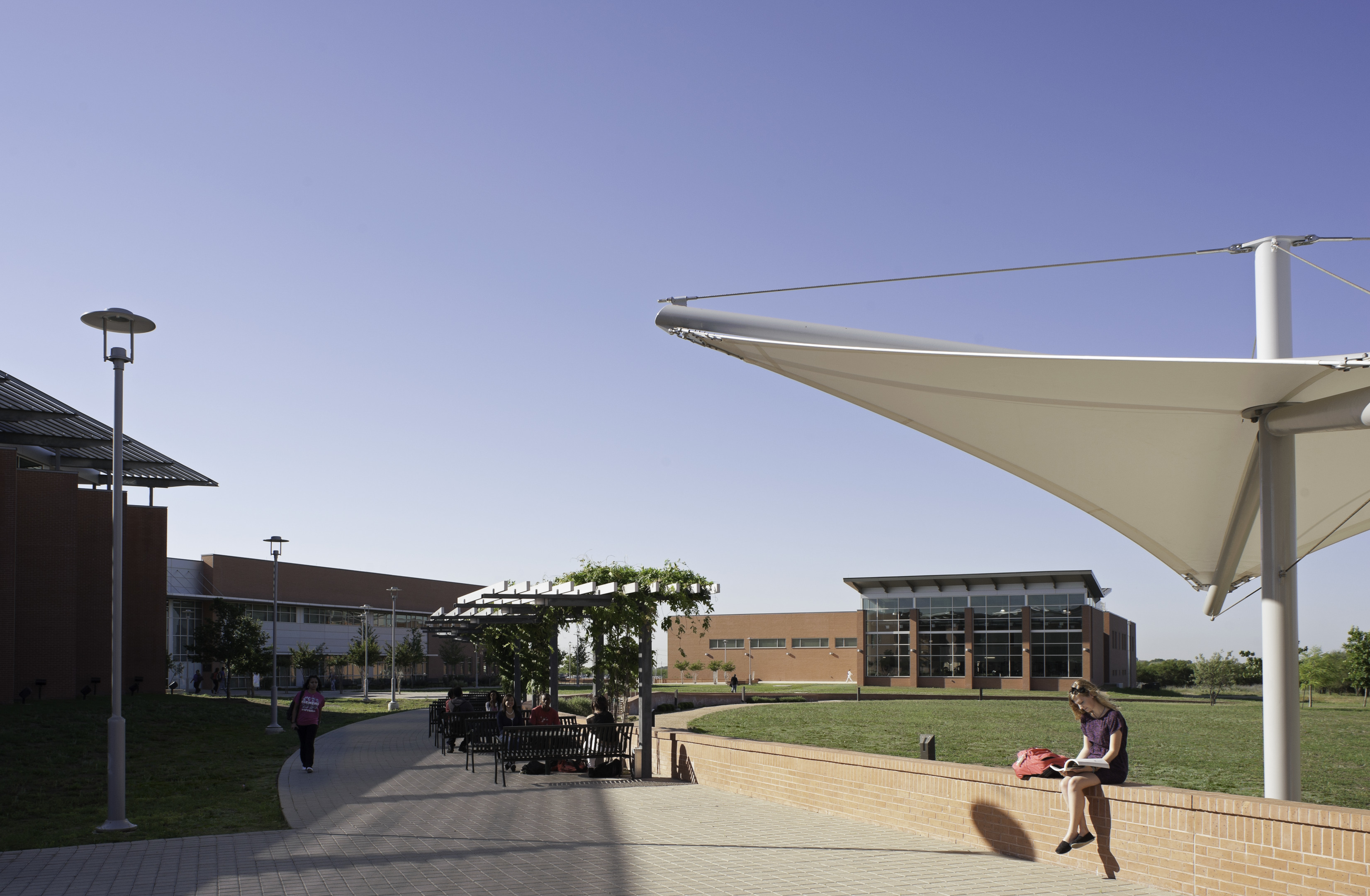 A person relaxes under a white canopy on a paved path at a modern campus, surrounded by several brick buildings and a clear blue sky, lost in thought as if orchestrating an auto draft of ideas.