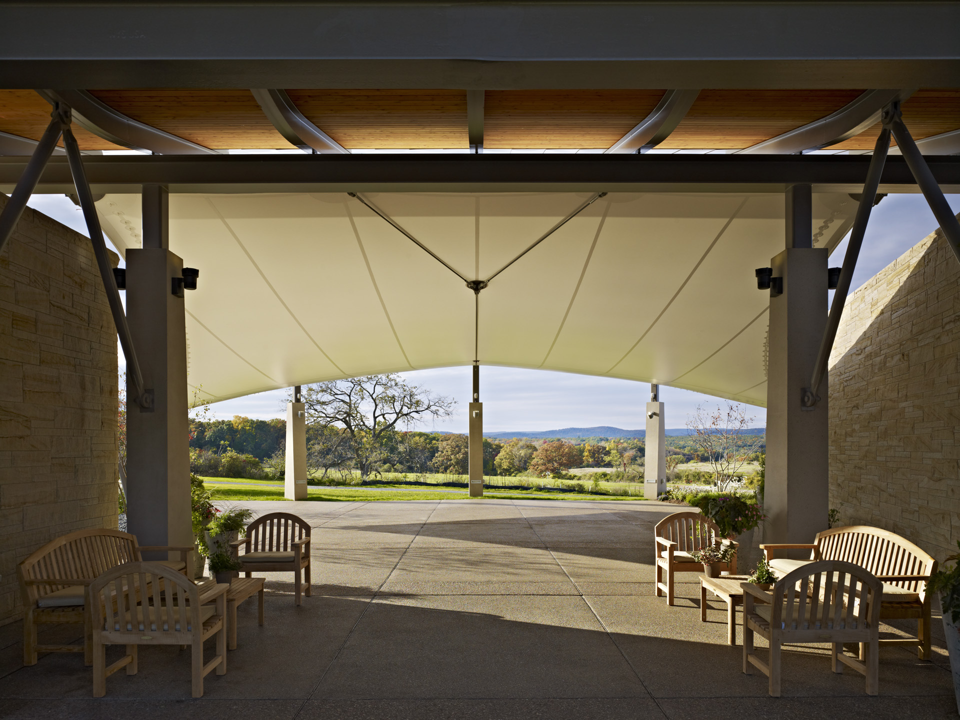 An open-air pavilion with a large canopy roof and wooden chairs provides an Auto Draft experience, overlooking a scenic landscape with trees and hills in the background.