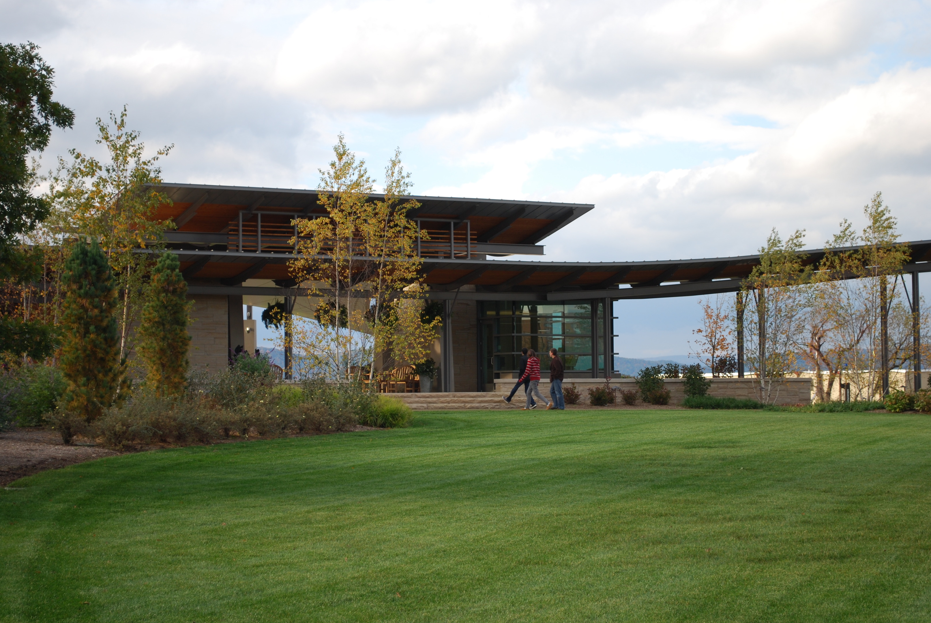 A modern building with large windows and a flat roof, resembling an architectural auto draft, is surrounded by trees and a well-maintained lawn. Two people walk on a path in the foreground under the moody, cloudy sky.