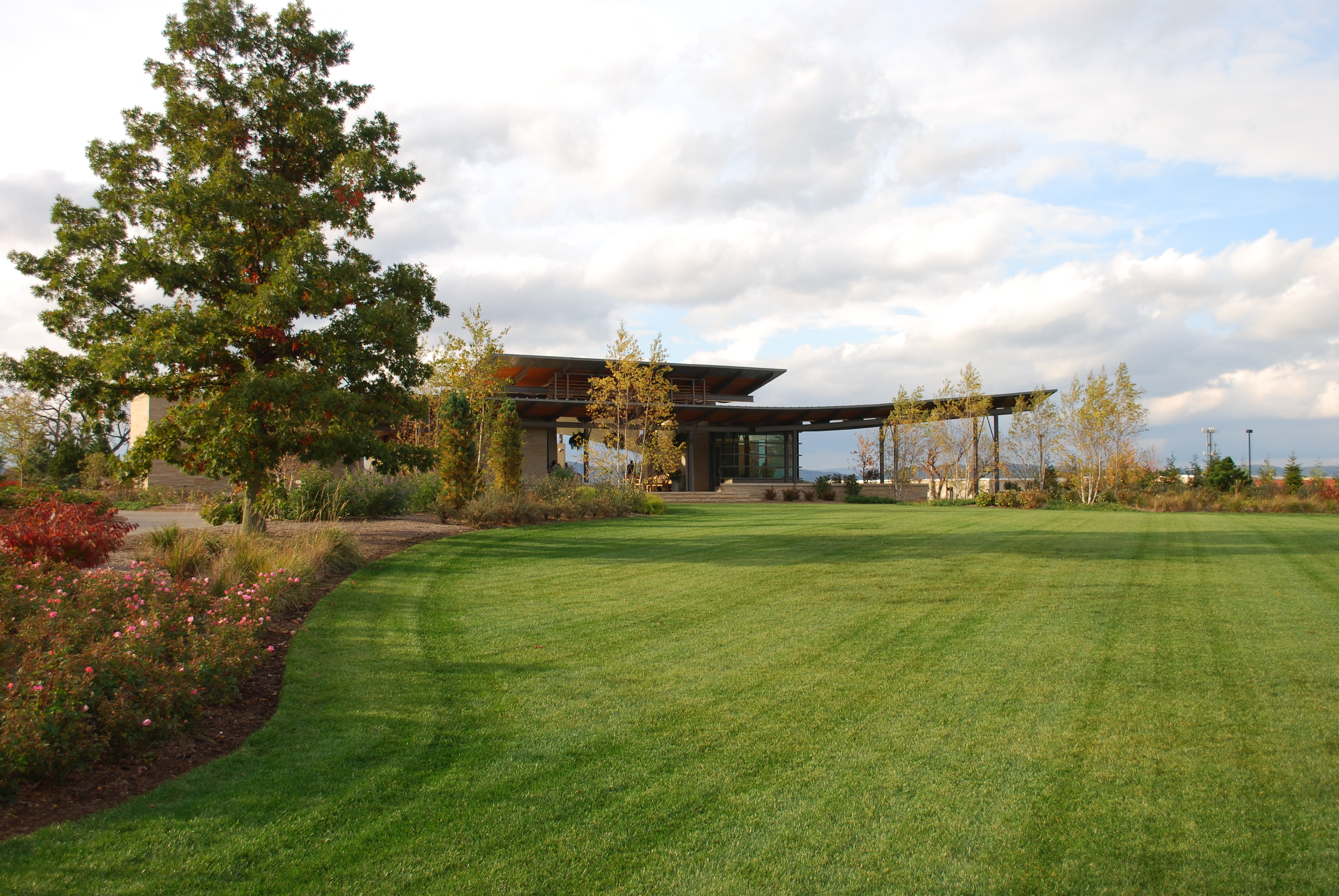 A modern building with a sloped roof stands elegantly amidst a sprawling, well-manicured lawn and towering trees, framed by a partly cloudy sky—a serene yet sophisticated setting ideal for capturing the essence of innovative architecture.