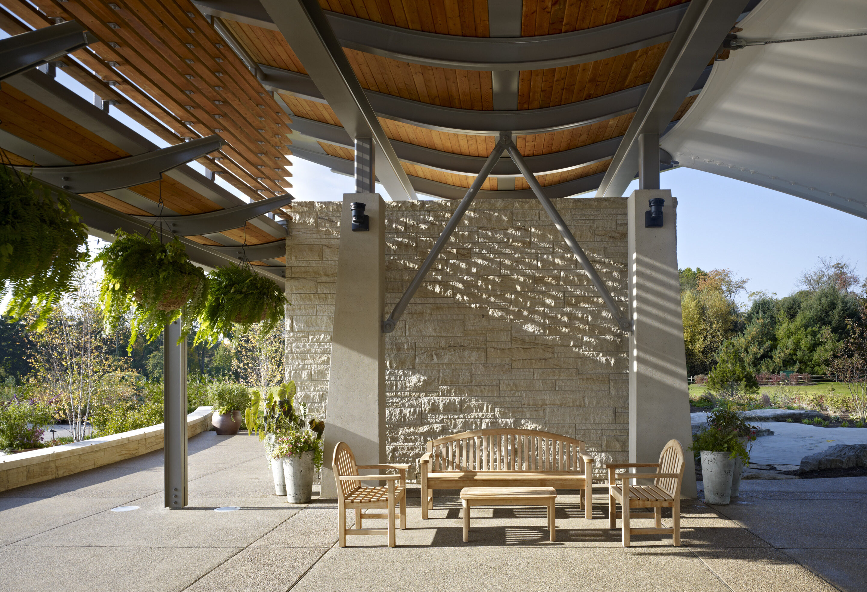 The modern outdoor seating area, reminiscent of an auto draft design, features wooden benches and chairs under a sleek curved metal and wood roof. Potted plants hang on the left, while a stone wall casts geometric shadows in the background.