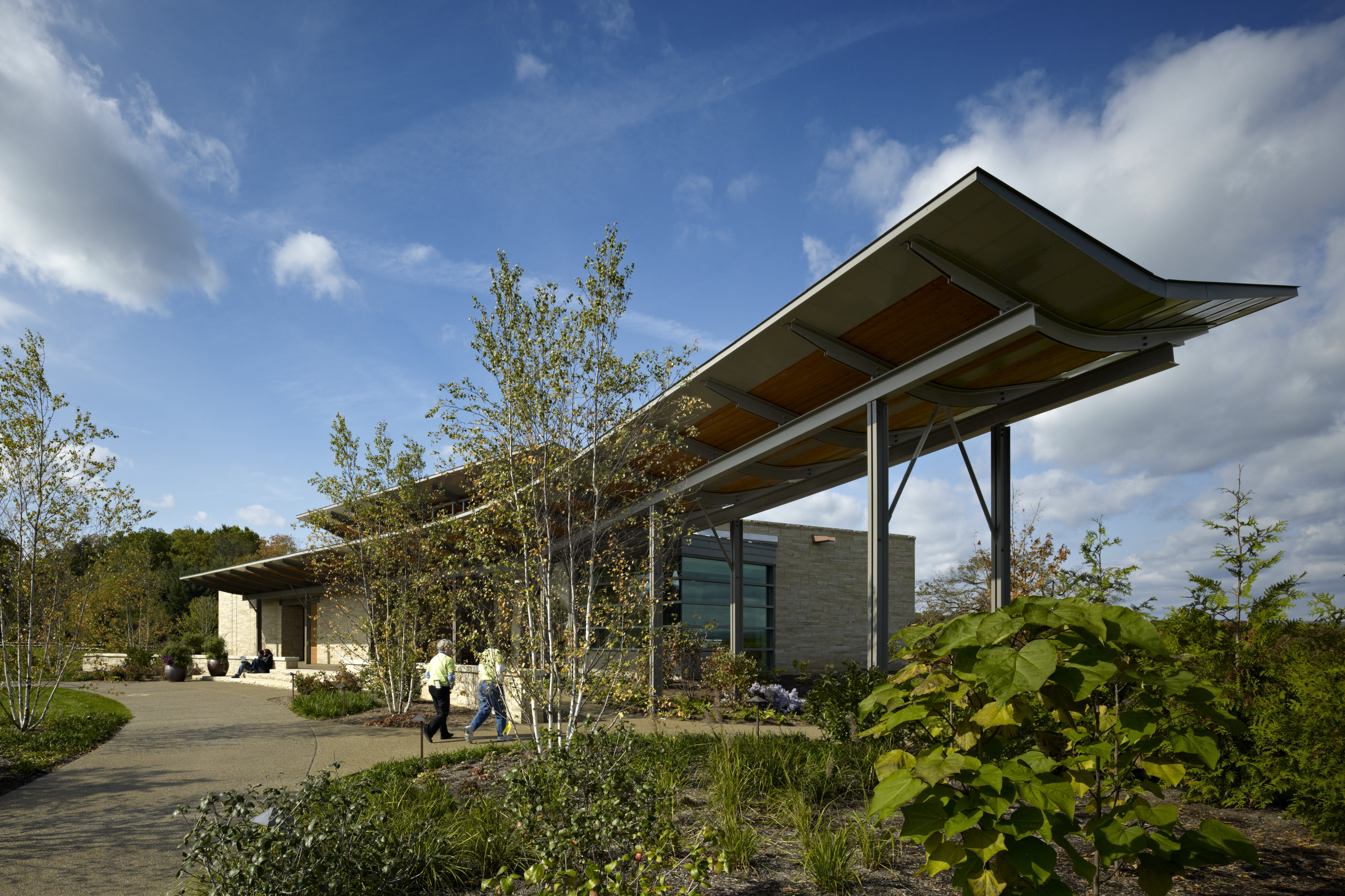 A modern building with an angled roof and glass facade stands surrounded by lush greenery, embodying an auto draft design. In the foreground, two people stroll along a winding path.