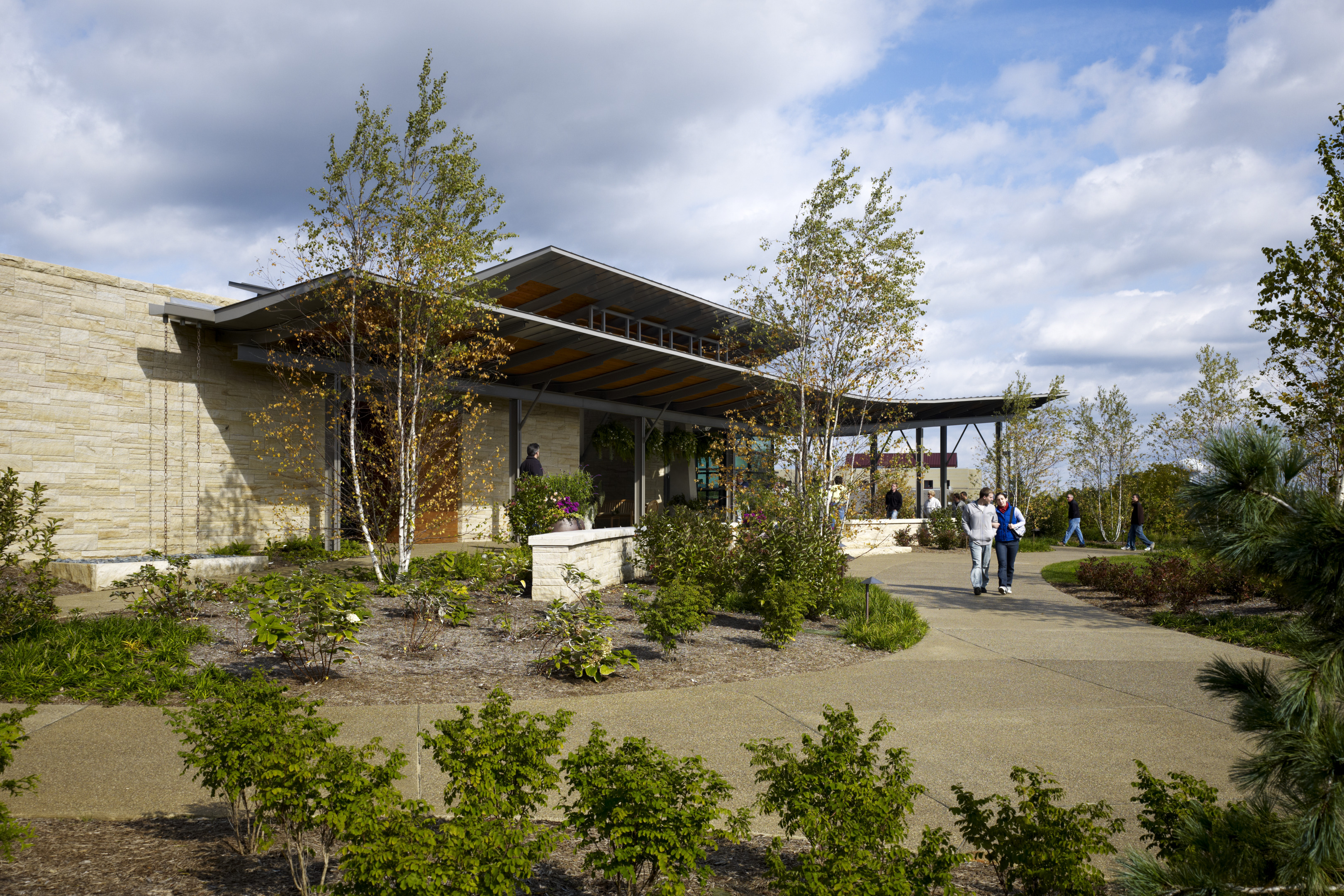 A modern building entrance with large windows and tall trees is surrounded by landscaped greenery. A few people walk along the pathway under a partly cloudy sky, while the scene seems to spring to life like an auto draft rendering of urban tranquility.