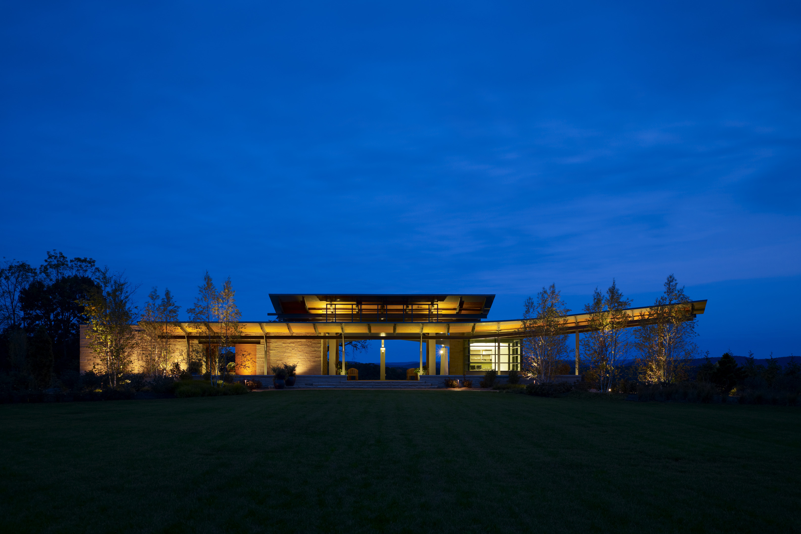 A modern building with expansive glass windows glows like a beacon under a deep blue sky, surrounded by trees and a neatly manicured lawn.