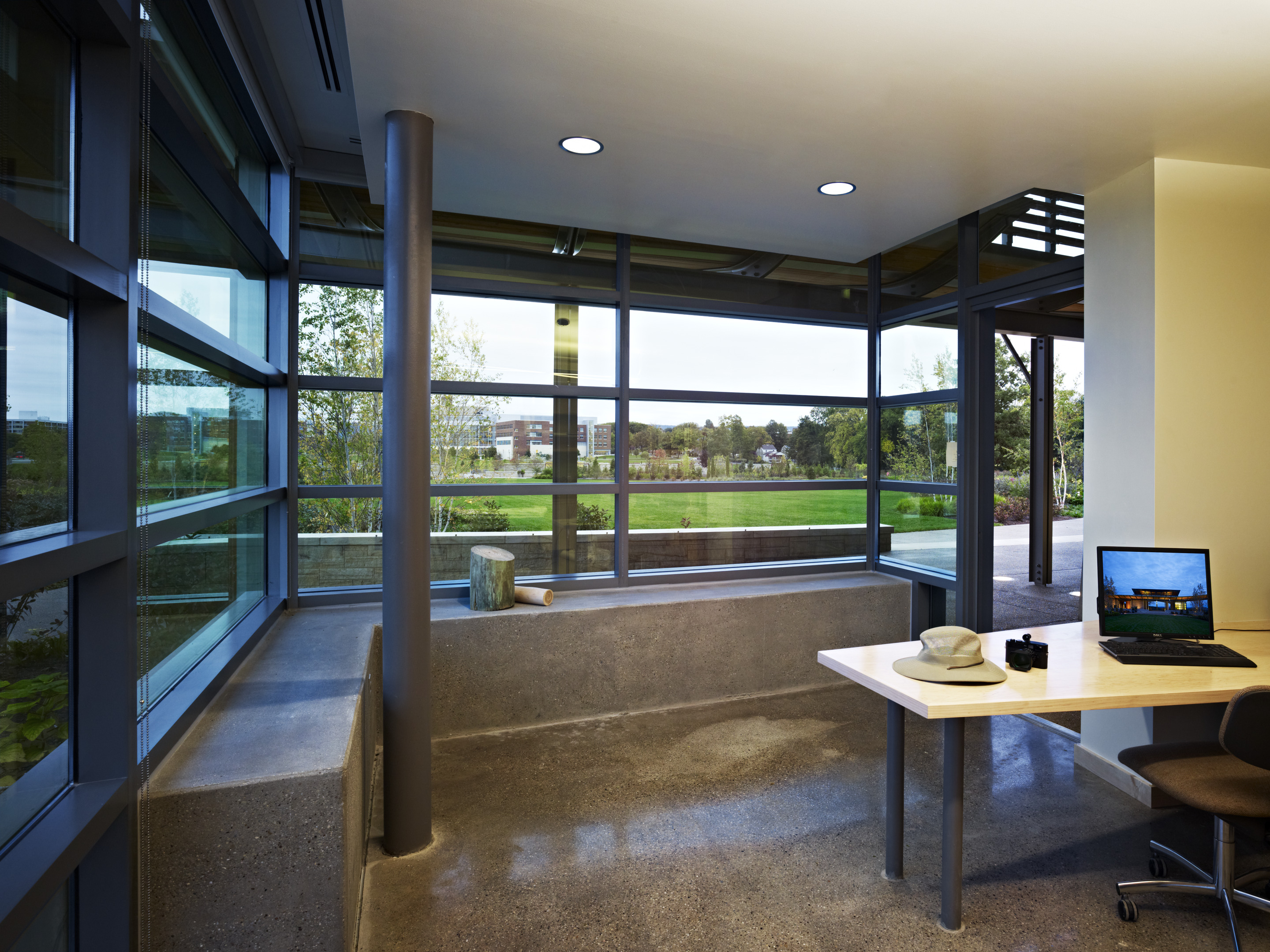 A modern office with large windows showcasing a lush outdoor view is the perfect setting for inspiration. Near the windows, a desk equipped with a laptop, hat, and camera awaits your next auto draft of creativity.