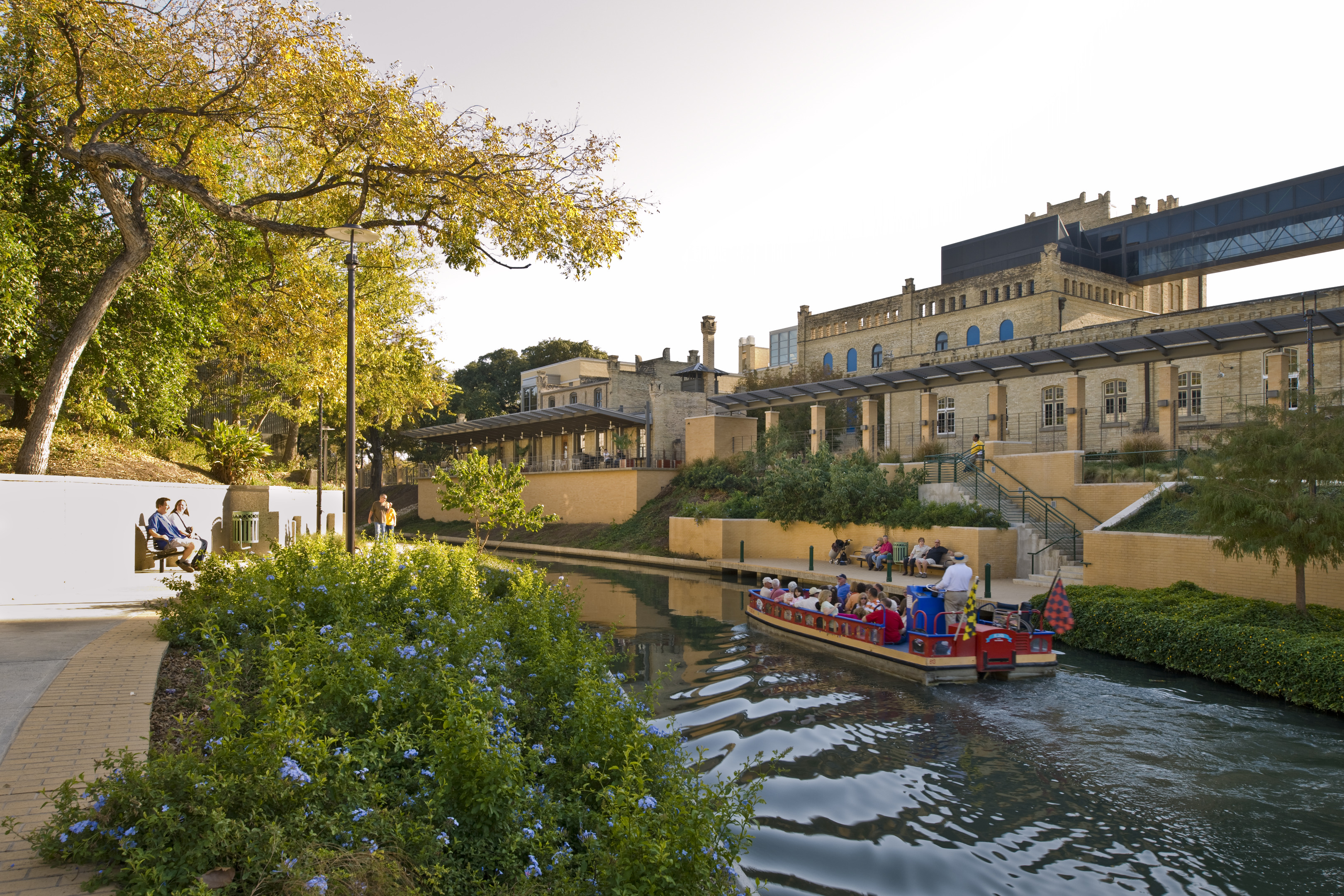 A canal with a boat carrying passengers glides past a historic building and lush greenery under a clear sky, while people relax on a bench by the water's edge, enjoying the serene view.