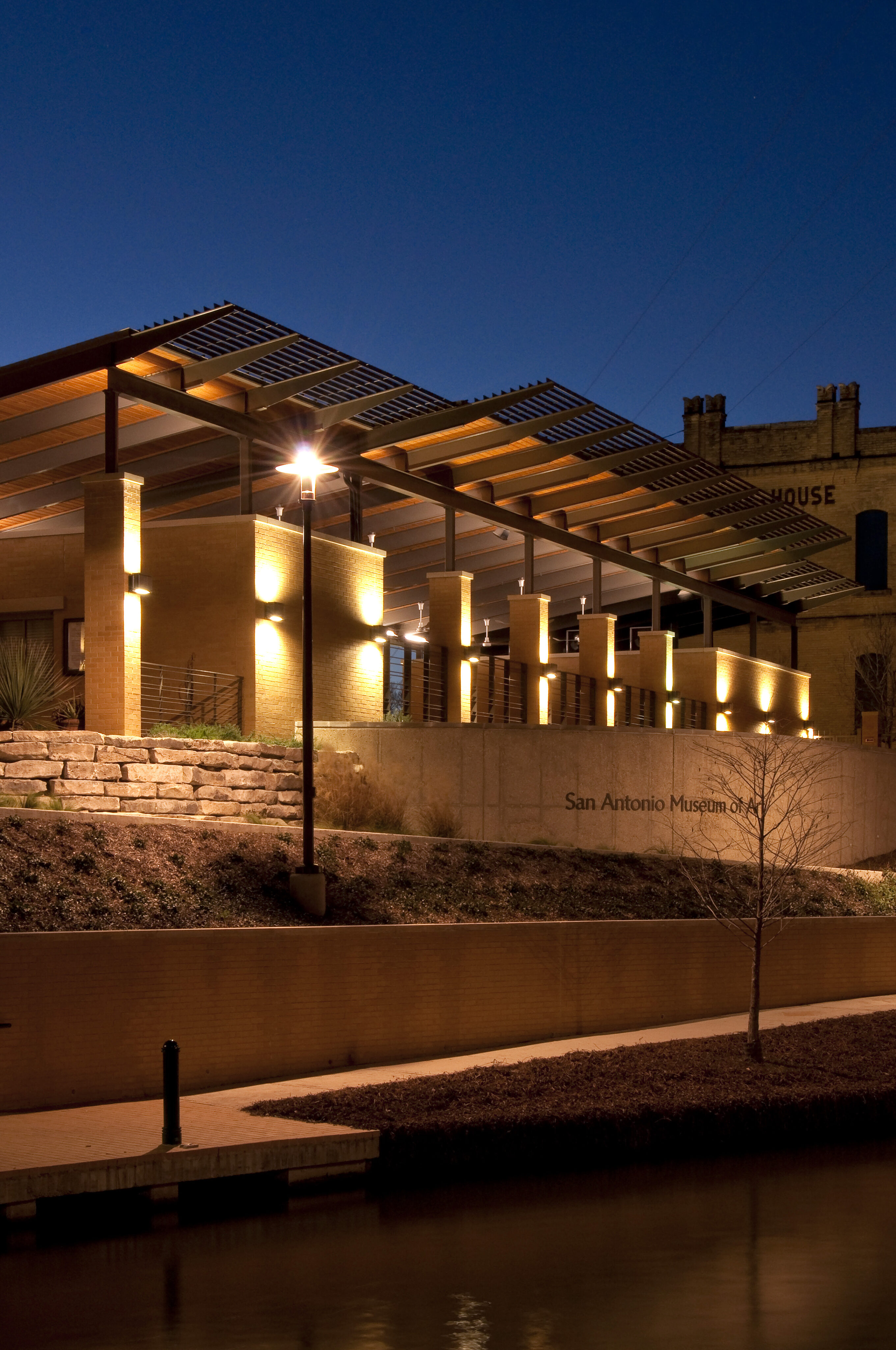 An outdoor view of a modern building with a lit façade and overhanging structure hovers beside a river at twilight, its reflection casting an almost auto-draft quality across the water.