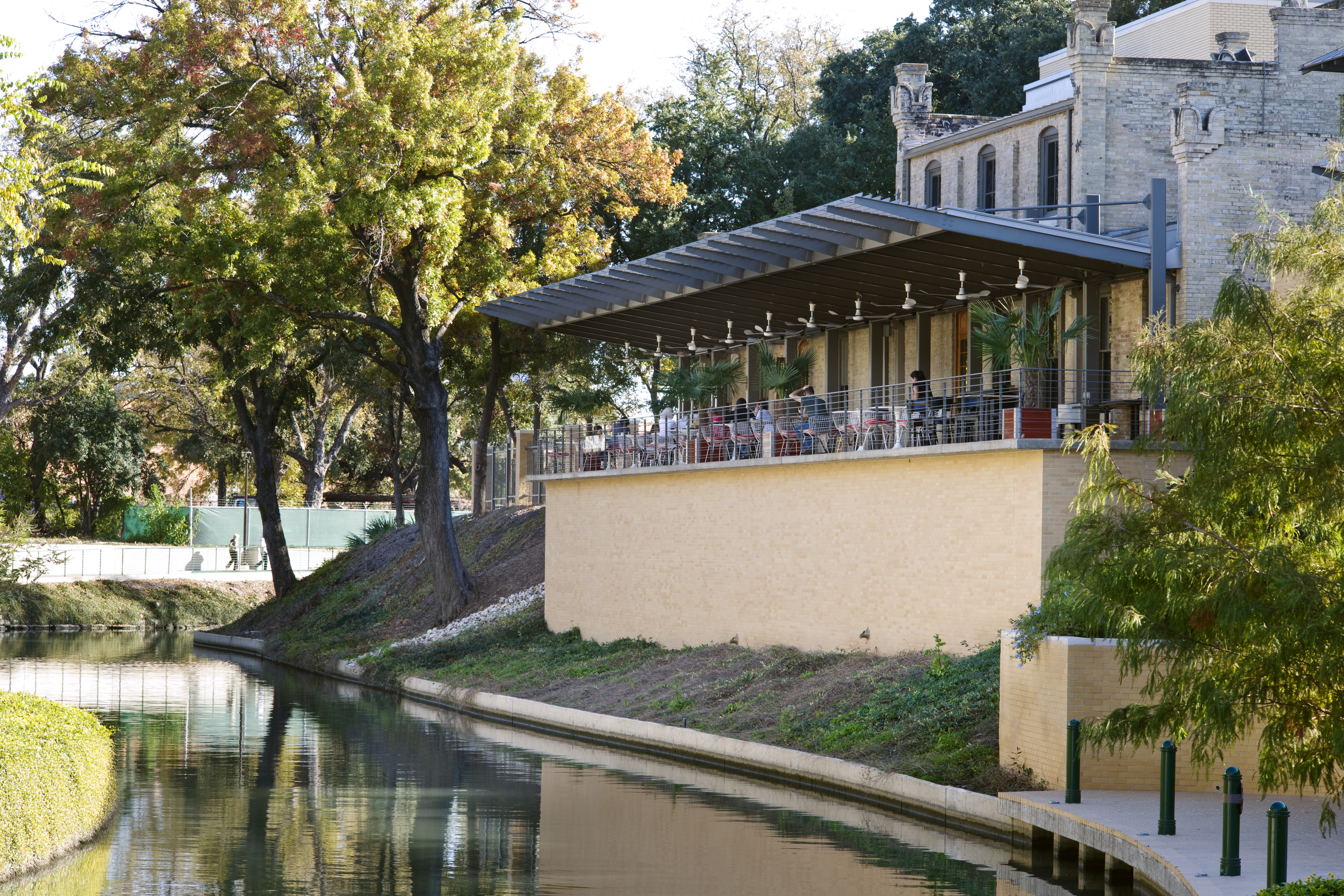 Outdoor restaurant with patrons on a covered terrace overlooking a tree-lined river on a sunny day, seamlessly blending into the historic stone building in the background. The captivating scene feels as if crafted by auto draft, effortlessly capturing nature and heritage in harmony.