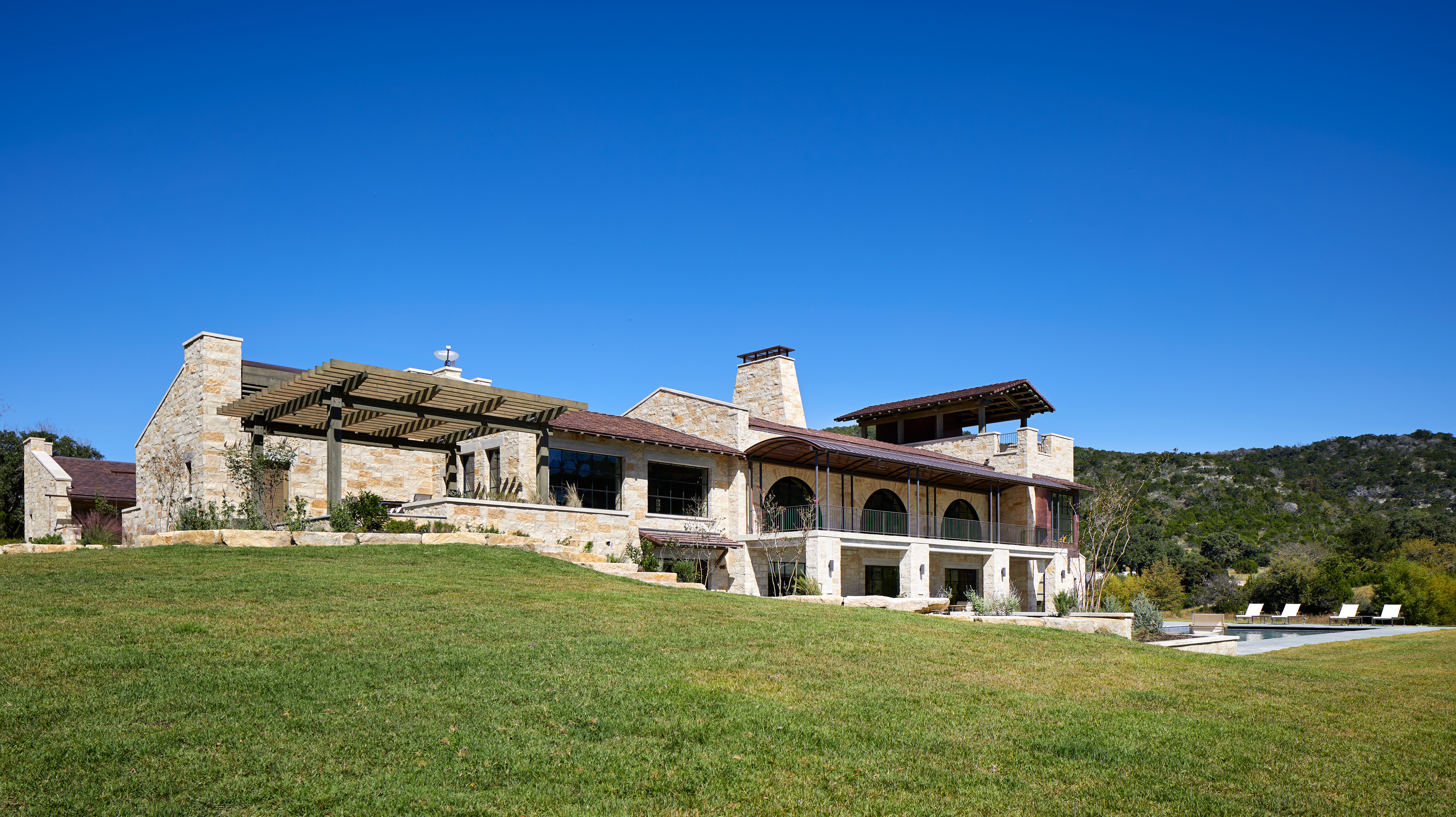 Nestled on a grassy hill under a clear blue sky, Skull Creek Ranch boasts a large stone house with a patio and pool in the backyard, offering the perfect blend of rustic charm and modern luxury.