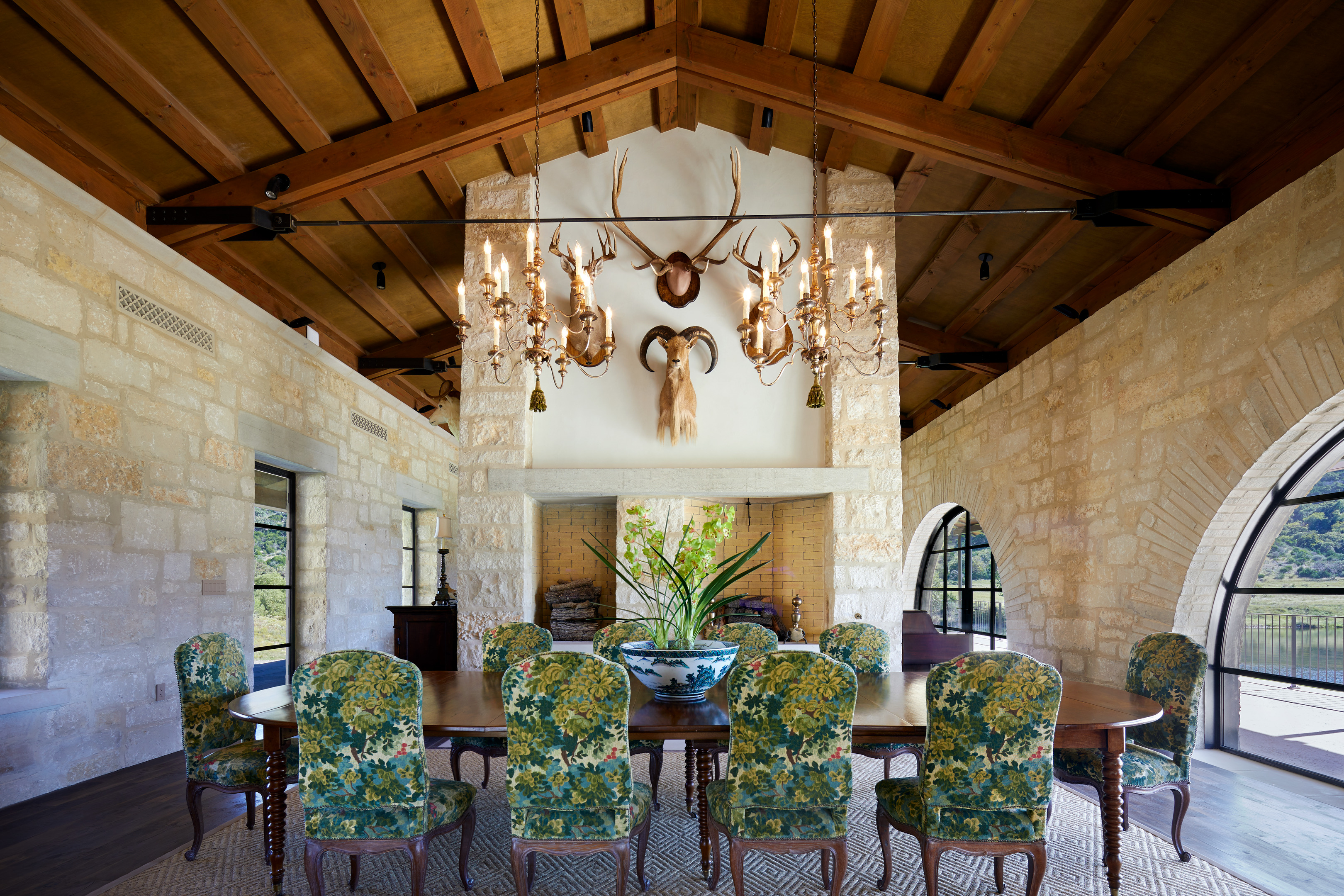 A rustic dining room at Skull Creek Ranch features a wooden ceiling, stone walls, and arched windows. A large table with floral chairs takes center stage, while chandeliers and animal mounts enhance the charm.