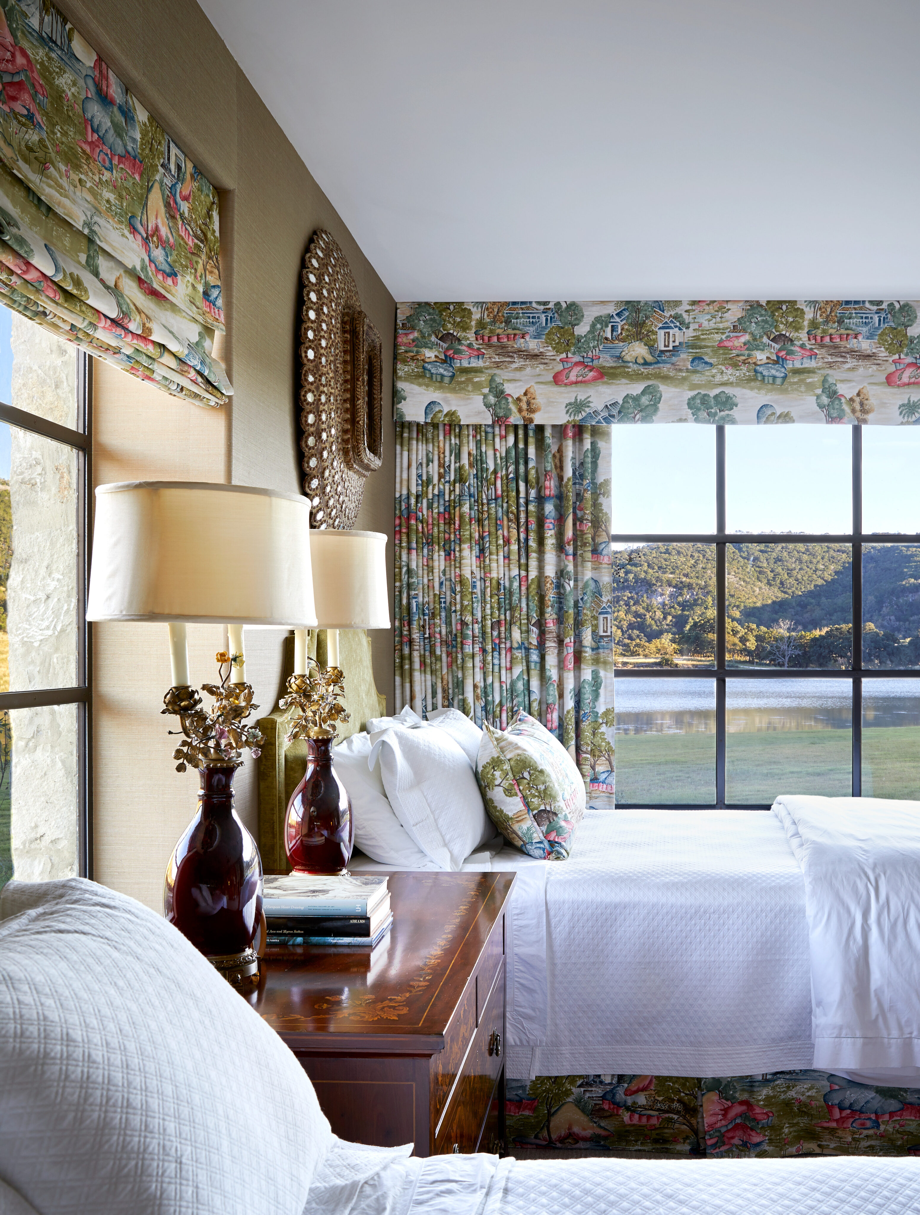 The bedroom at Skull Creek Ranch features two beds, complemented by floral curtains and large windows that offer a scenic view. A wooden side table supports two lamps and decorative items, adding charm to this serene retreat.