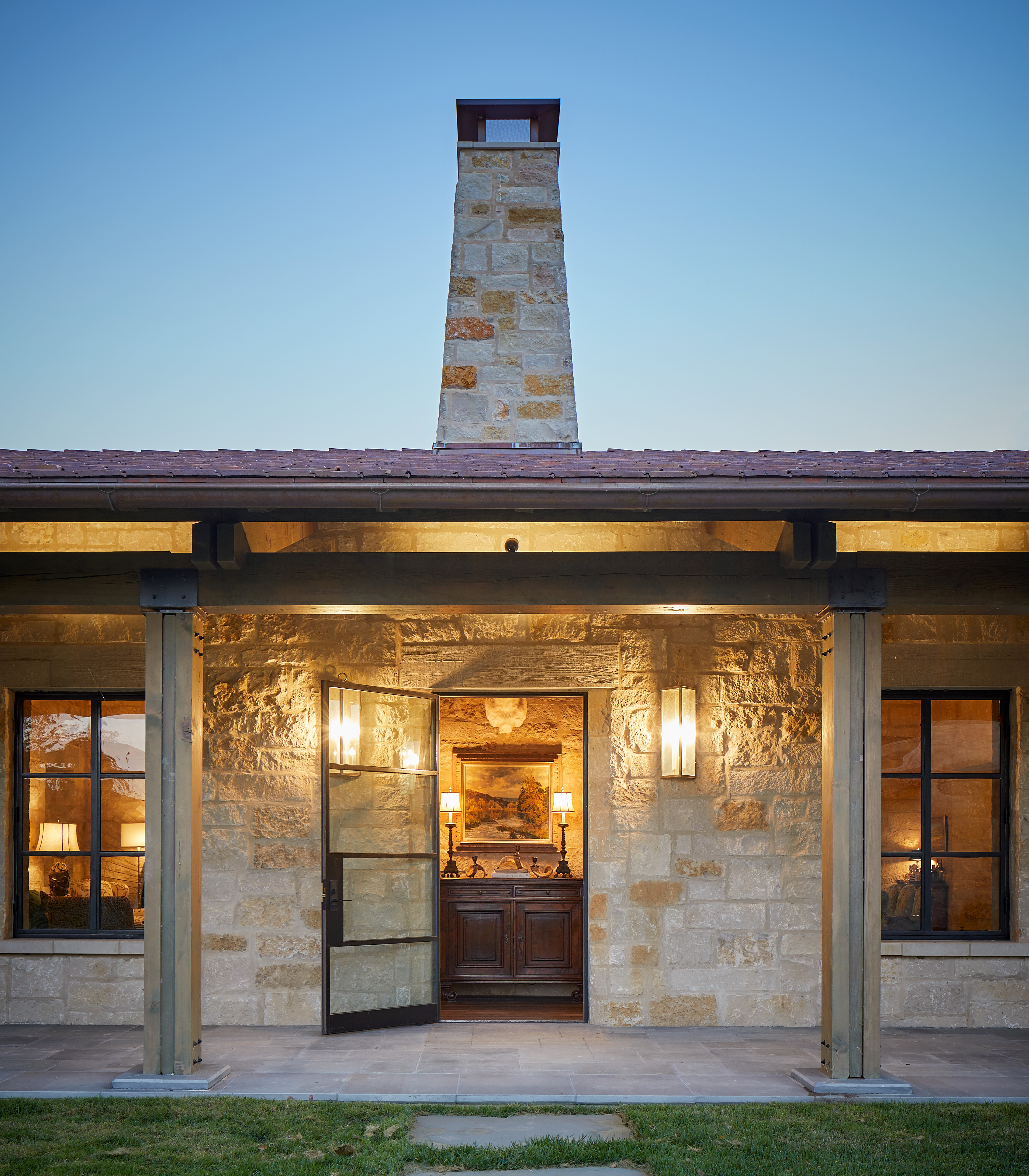 A stone building facade at Skull Creek Ranch features a central chimney, large glass doors, and lit lanterns on either side. The interior appears warmly lit and inviting.