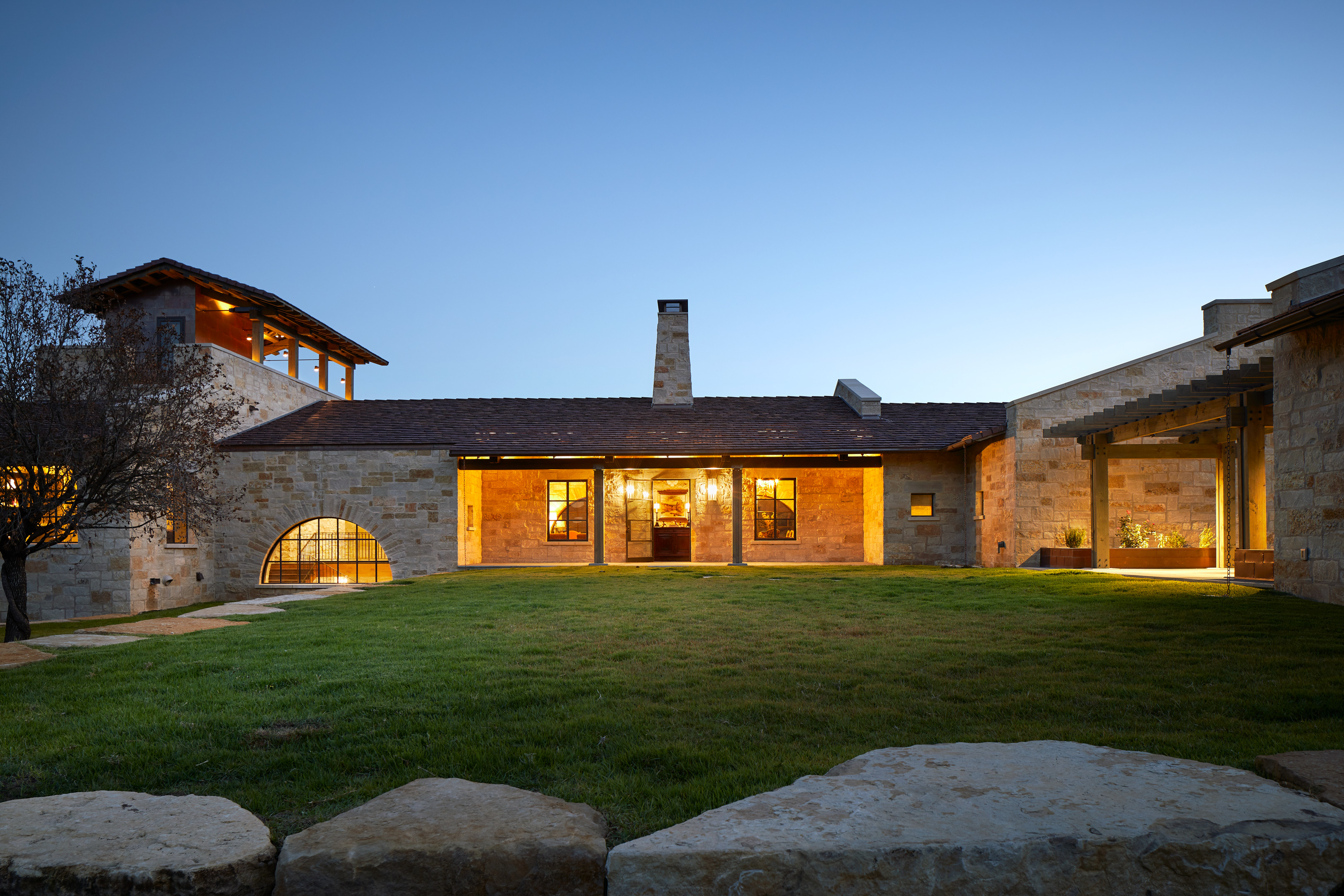 A stone house with warm interior lighting, a central chimney, and a unique tower feature stands proudly at Skull Creek Ranch against a twilight sky. In the foreground, a grassy lawn is framed by elegant stone borders.