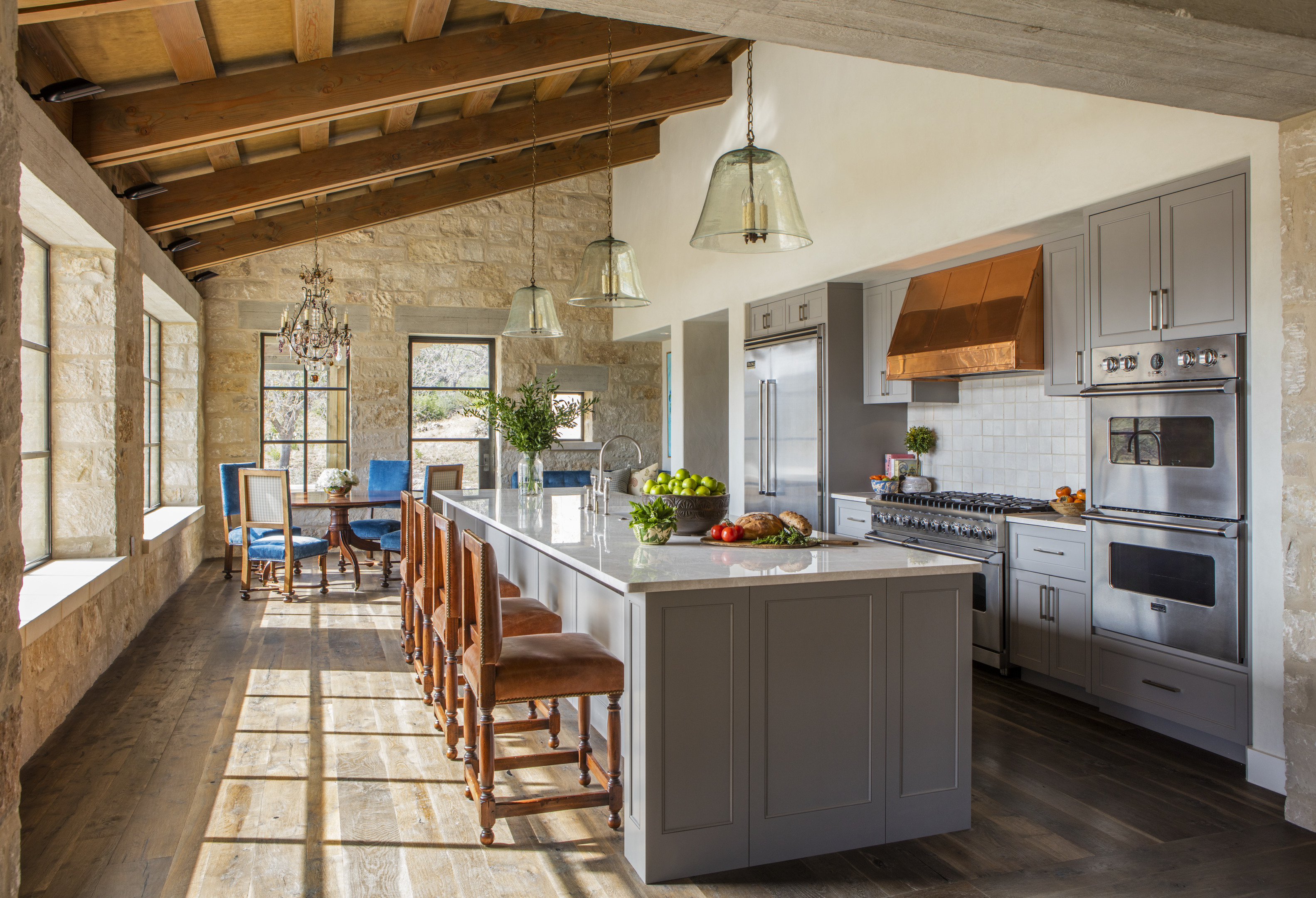 The sunlit kitchen at Skull Creek Ranch features wooden beams, a marble-topped island, and gray cabinetry. Stainless steel appliances complement the rustic charm, while glass pendant lights hang gracefully above the island with chairs.