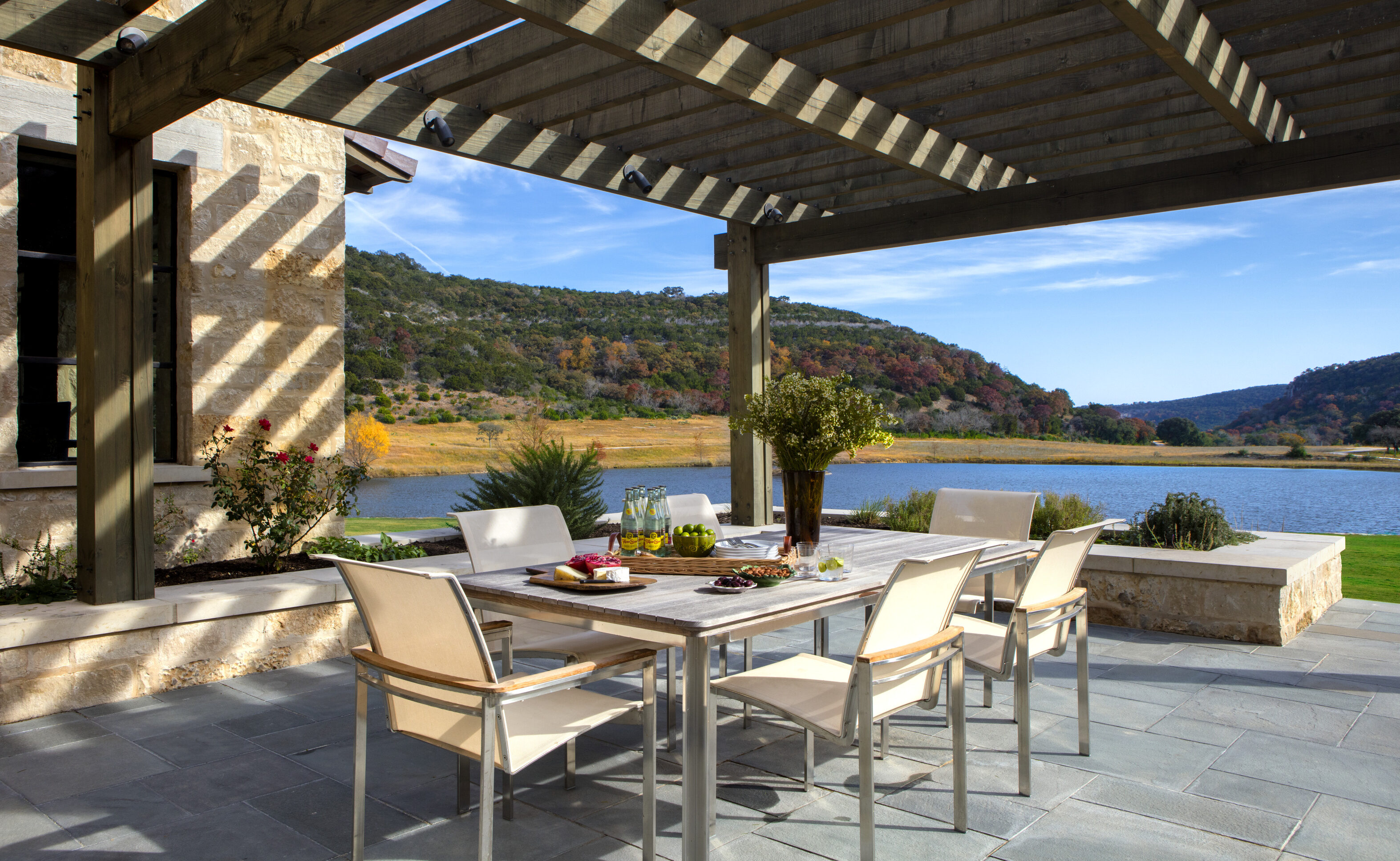 The outdoor patio at Skull Creek Ranch features a wooden pergola, a dining table set for four, and a scenic view of the lake and hills in the background.